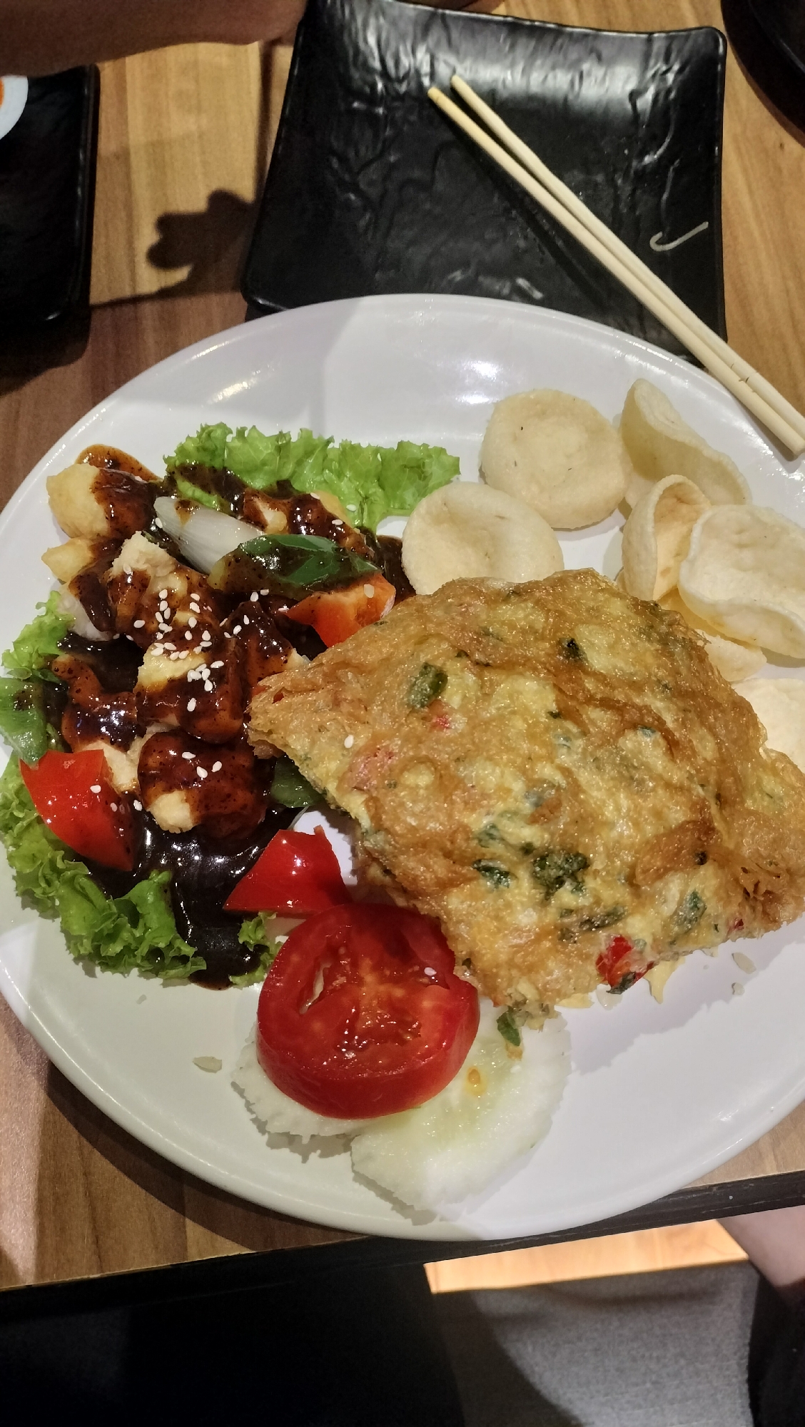 The image shows a close-up of a plate of food on a wooden table. The plate is white and round, filled with various food items. On the left side of the plate, there is a bed of lettuce topped with cubed pieces of food, possibly fried tofu or chicken, drizzled with a dark sauce and sprinkled with sesame seeds. Sliced tomatoes and green peppers are also visible among the lettuce and cubed items. To the right of this section, there is a golden-brown omelet filled with green herbs and red pieces, likely chili or bell pepper. Behind the omelet, there are several light-colored, circular crackers and what appear to be small, puffy crackers or chips. At the bottom of the plate, there is a slice of tomato placed on top of a slice of cucumber.
In the background, a black rectangular plate with a textured surface is visible, holding a pair of wooden chopsticks. The setting appears to be a restaurant or dining area, suggested by the wooden table and the presence of plates and chopsticks. The lighting suggests it is daytime, possibly indoors. The focus is primarily on the food, creating an appetizing presentation. There is no visible text in the image.