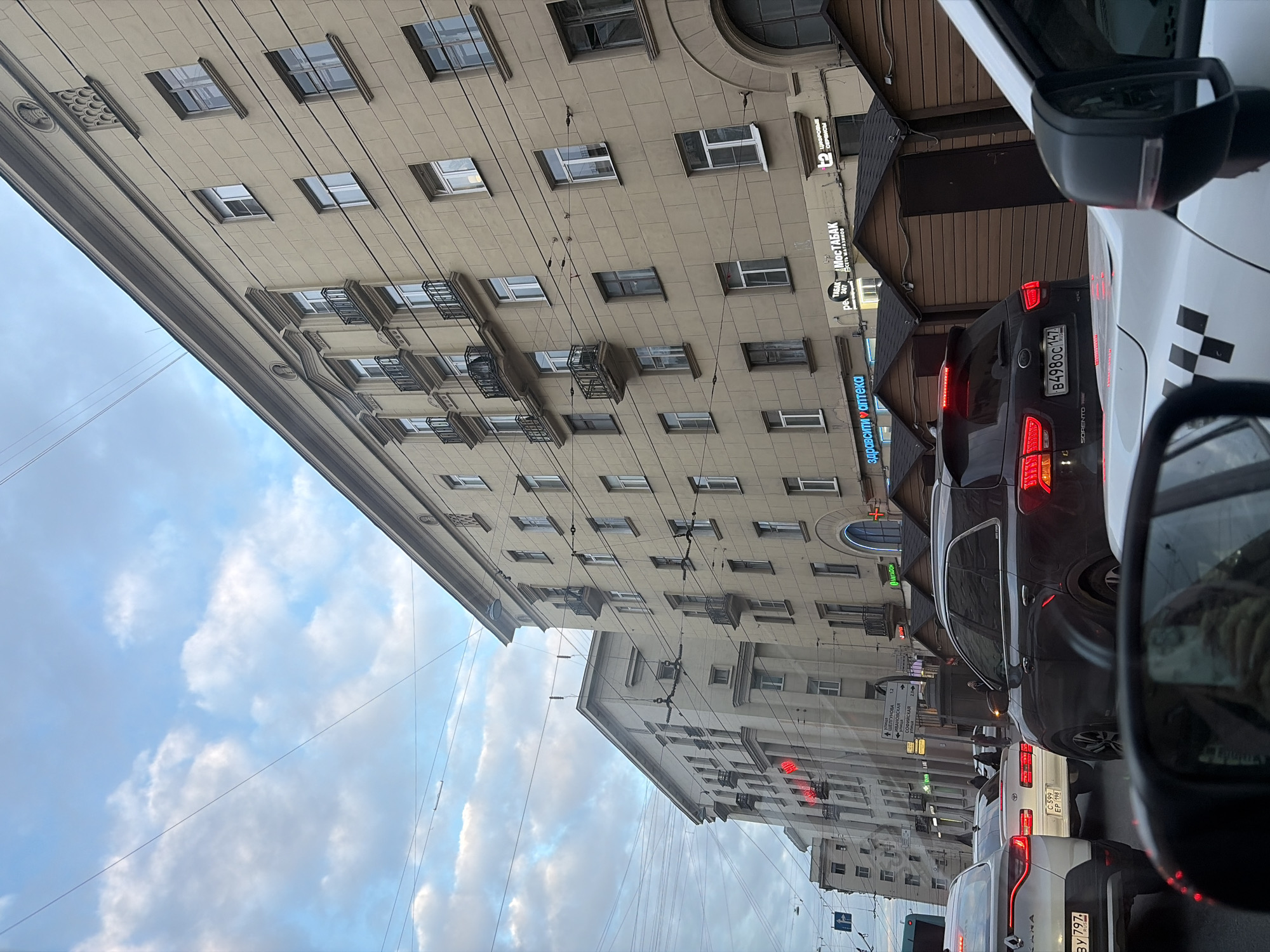 The image captures a street scene in Saint Petersburg, Russia. It appears to be taken from within a car, looking out onto a street with buildings on either side. The building on the left is tall and multi-storied, with numerous windows and some small balconies. There are many overhead wires running across the sky, which is filled with clouds.
On the right side of the image, there are cars in a traffic jam. There is a grey Kia Sorento with license plate B498CC 147 and a white taxi vehicle in the lane next to the car the photo was taken in. The taxi has checkered markings along its side. Various storefront signs in Russian are visible, including "МосТАБАК," "здравсити аптека," and "МЕГАФОН". There are other street signs indicating directions to "ШЕЛГУНОВА," "ИВАНОВСКАЯ," and "СОФИЙСКАЯ".