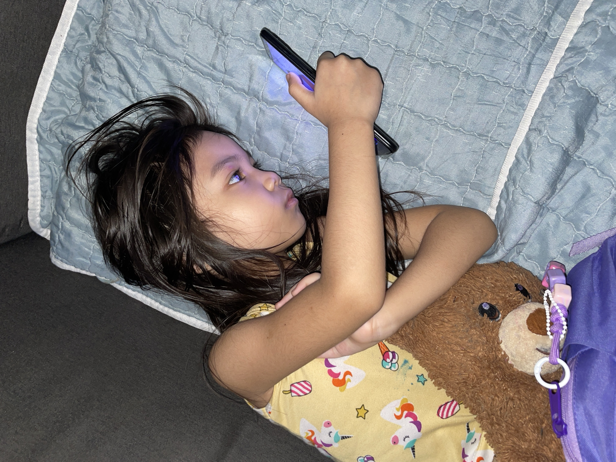 A young Filipino girl lies on her back on a quilted blue blanket, her head slightly turned to her left. She is holding a smartphone in her right hand, angled towards her face, and appears to be engrossed in its screen, which emits a bright blue light. Her dark hair is spread around her head. She is wearing a yellow t-shirt with a pattern of unicorns and ice cream cones. To her right, a brown stuffed bear is visible, partially covered by a purple object, possibly a backpack or a pillow. The girl's arms are crossed over her chest. The scene suggests a moment of personal entertainment or relaxation, likely indoors given the lighting. The location is specified as Davao City, Philippines. There is no visible text in the image.