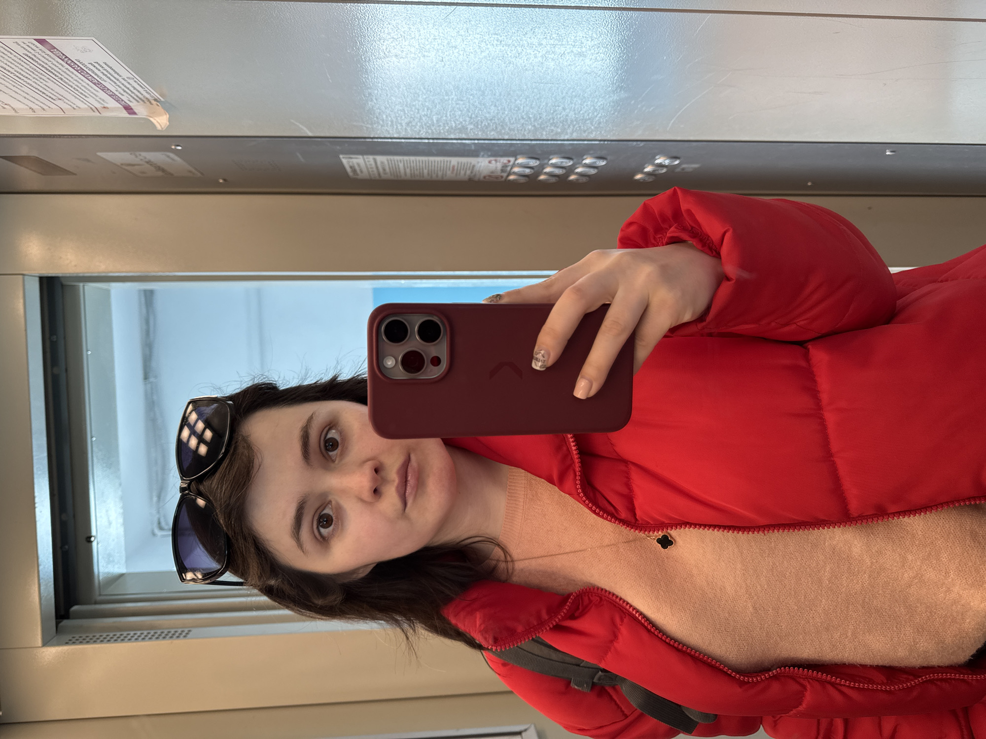 An elevated full-body self-portrait shows a young adult female inside an elevator in Izhevsk, Russia. The woman, with dark brown hair and light skin, wears a red puffer jacket over an orange-peach knit sweater, a gold-colored necklace with a black four-leaf clover pendant, and a dark-colored backpack strap over her left shoulder. Black sunglasses rest on top of her head. She holds a dark red smartphone, featuring a triple camera array, in her right hand, extended to capture the image. Her fingernails have a light-colored, patterned polish. The elevator interior consists of light beige walls and a reflective panel on the right. This panel displays an elevator control interface with multiple silver-colored buttons arranged vertically and a paper sign taped above them. The sign contains Cyrillic text, including "ИЖЕВСК" (Izhevsk), confirming the location. A ventilation grill is visible on the left wall. The lighting within the elevator is bright, casting reflections on the phone screen and sunglasses.