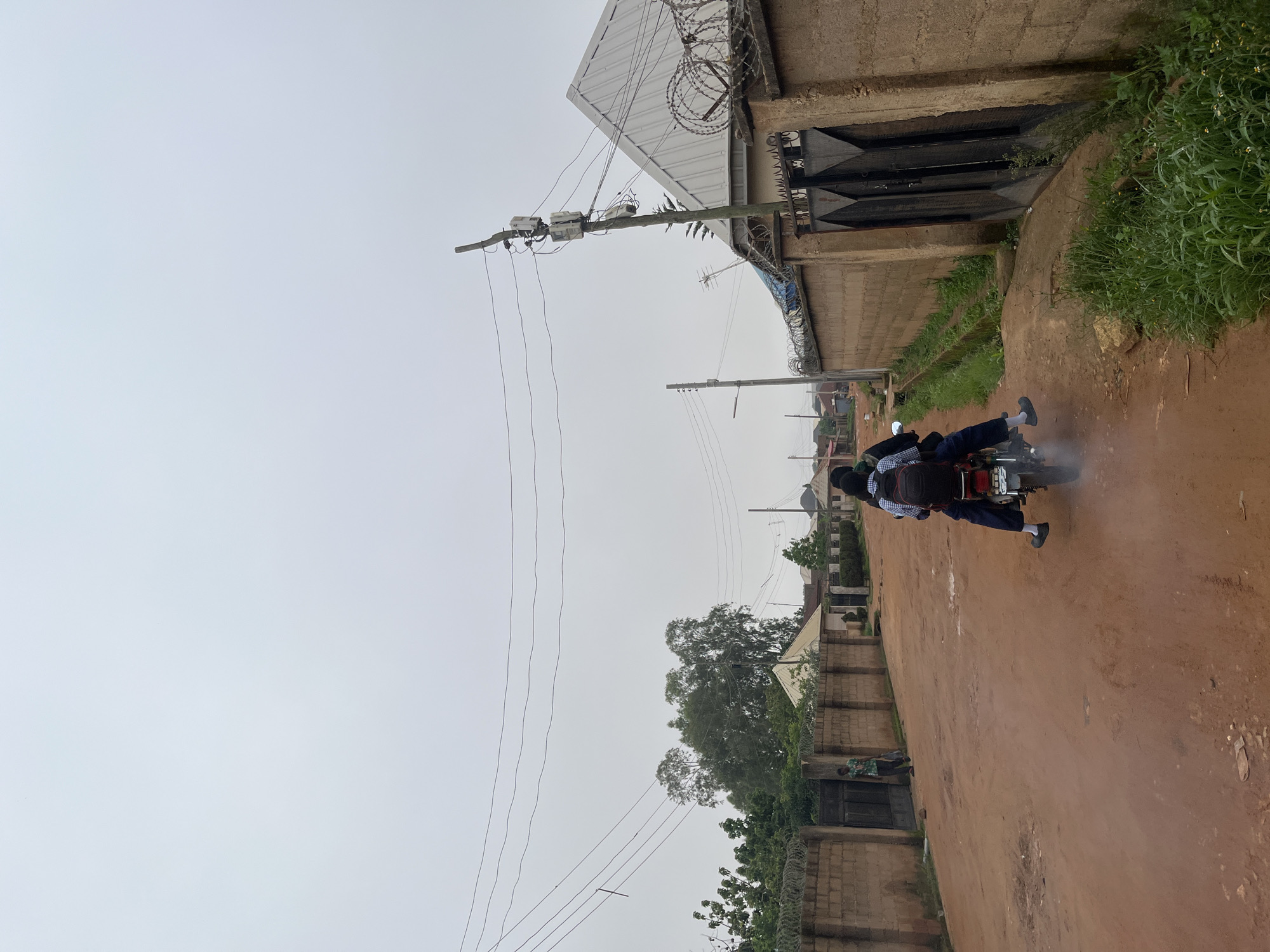 The image shows a dusty, reddish-brown dirt road in Jos, Nigeria, under an overcast sky. Two people, likely students given their school uniforms, are riding on a motorcycle. The rider in front wears a blue and white checkered shirt and dark pants, while the passenger behind wears a similar uniform and a black backpack. They are moving away from the viewer, kicking up a small amount of dust.
To the right of the road, there is a tall, weathered brick wall topped with barbed wire, indicating a secure property. Behind the wall, green vegetation thrives. Further along, a large, dark metal gate is visible, set within a concrete structure. The building adjacent to the gate has a corrugated metal roof and more barbed wire.
On the left side of the road, a utility pole with electrical meters and wires stretches upwards, disappearing into the sky. Additional utility poles and wires are visible in the background. The scene suggests a residential or commercial area on the outskirts of a town, characterized by unpaved roads and functional, somewhat utilitarian structures. The overall atmosphere is muted due to the cloudy weather, suggesting it might be early morning or late afternoon, or simply a day with little sunshine. There is no visible text in the image.