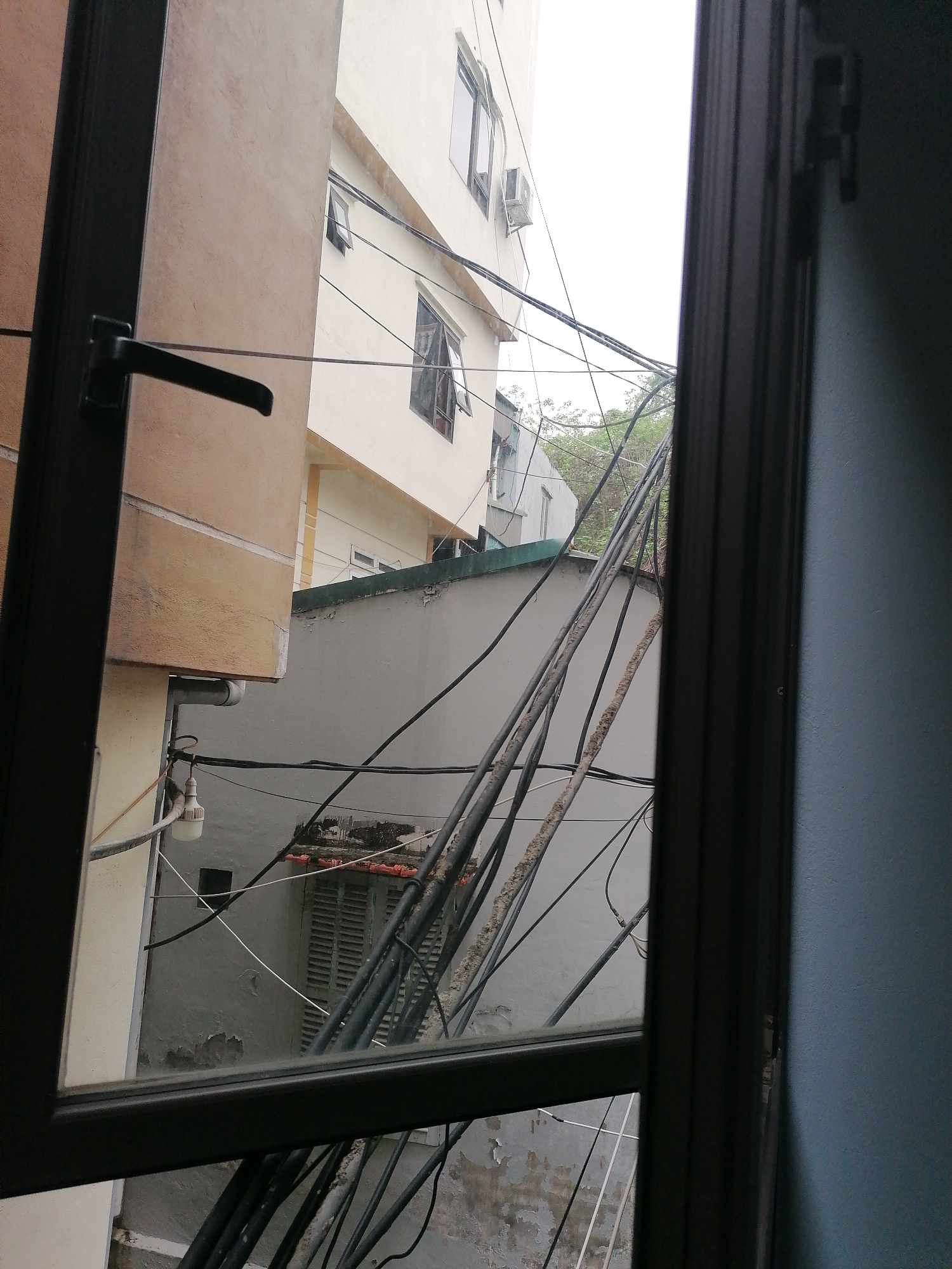 The image captures a view looking out of a window in an urban environment, likely in Bai Chay Ward, Vietnam. The dark frame of the window and a black handle are prominent in the foreground, with a segment of a blue-painted wall visible on the far right.
Beyond the window, a dense and complex network of overhead electrical or communication cables forms a chaotic web, crisscrossing the scene. Many of these cables appear thick and show signs of wear and tear, with some exhibiting white, crusty residue.
Several buildings are visible. To the immediate left is a beige or light brown building, appearing close to the window. Further back and dominating the upper central part of the view is a multi-story white building with several windows, some of which are open, and an outdoor air conditioning unit. Below this, a gray building or wall features a closed slatted window or shutter and displays noticeable peeling paint, indicating age and weathering. A suspended, white LED-style light bulb is attached to a fixture on the side of the beige building, near some pipes. In the background, beyond the gray structure and the tangle of wires, glimpses of green foliage suggest trees, and another lighter-colored building with a dark green roofline can be seen.
The sky appears overcast and bright, suggesting a cloudy day. There are no people visible, and no specific activity is taking place. The dense, unregulated mass of overhead wires is a common characteristic of urban landscapes in many Southeast Asian countries, consistent with the provided location context. No discernible text is visible in the image.