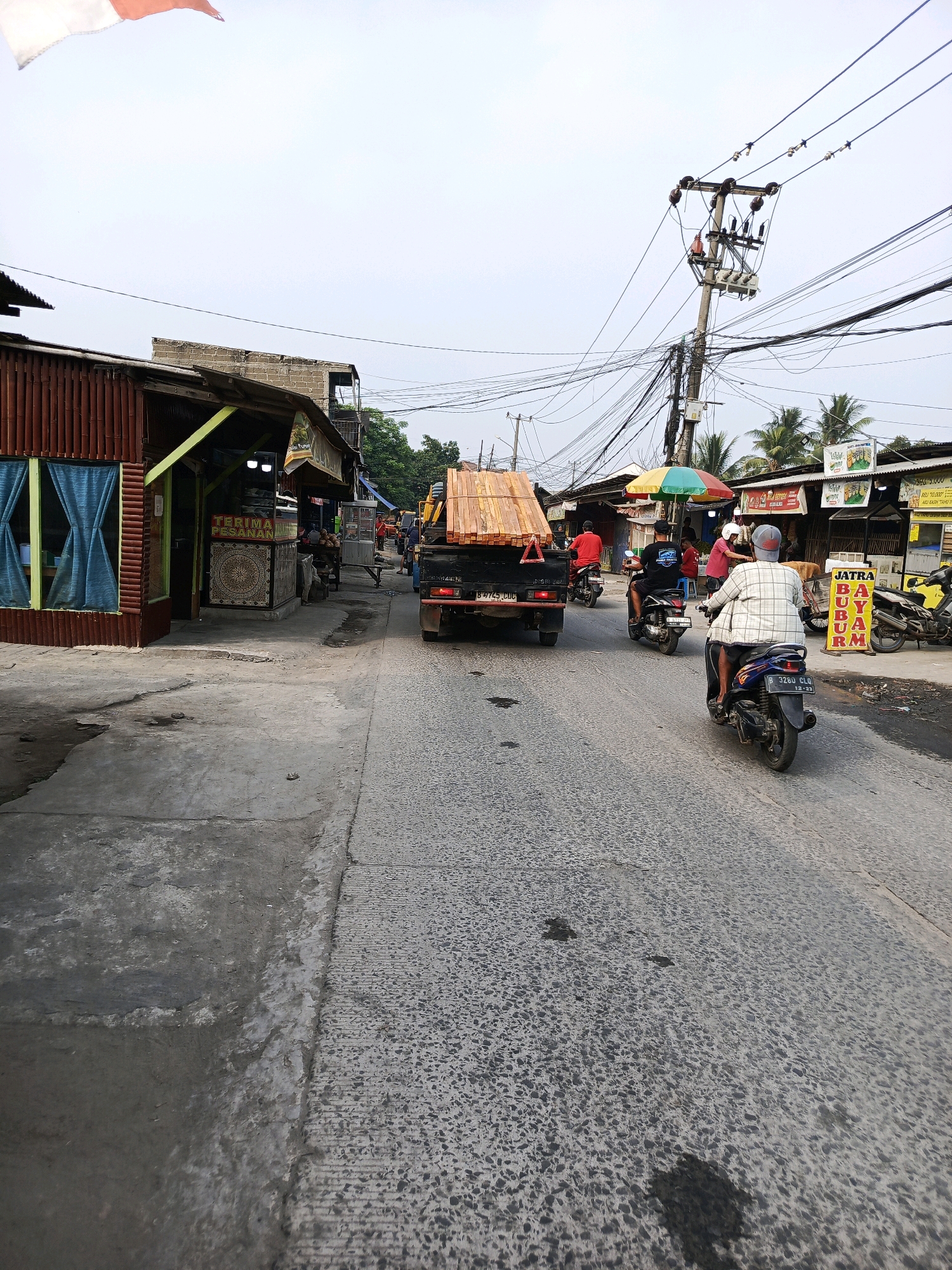 This is a street scene in Periuk, Indonesia. A black flatbed truck carrying a load of lumber is traveling down the road. To the left of the road are several buildings with corrugated metal roofs and wooden siding, one of which has a sign that reads "TERIMA PESANAN" (Accepting Orders). Further down the road, there is a power pole with numerous electrical wires and transformers. Several motorcycles are also visible, some with riders. The road surface is asphalt with some water stains. The sky is overcast.