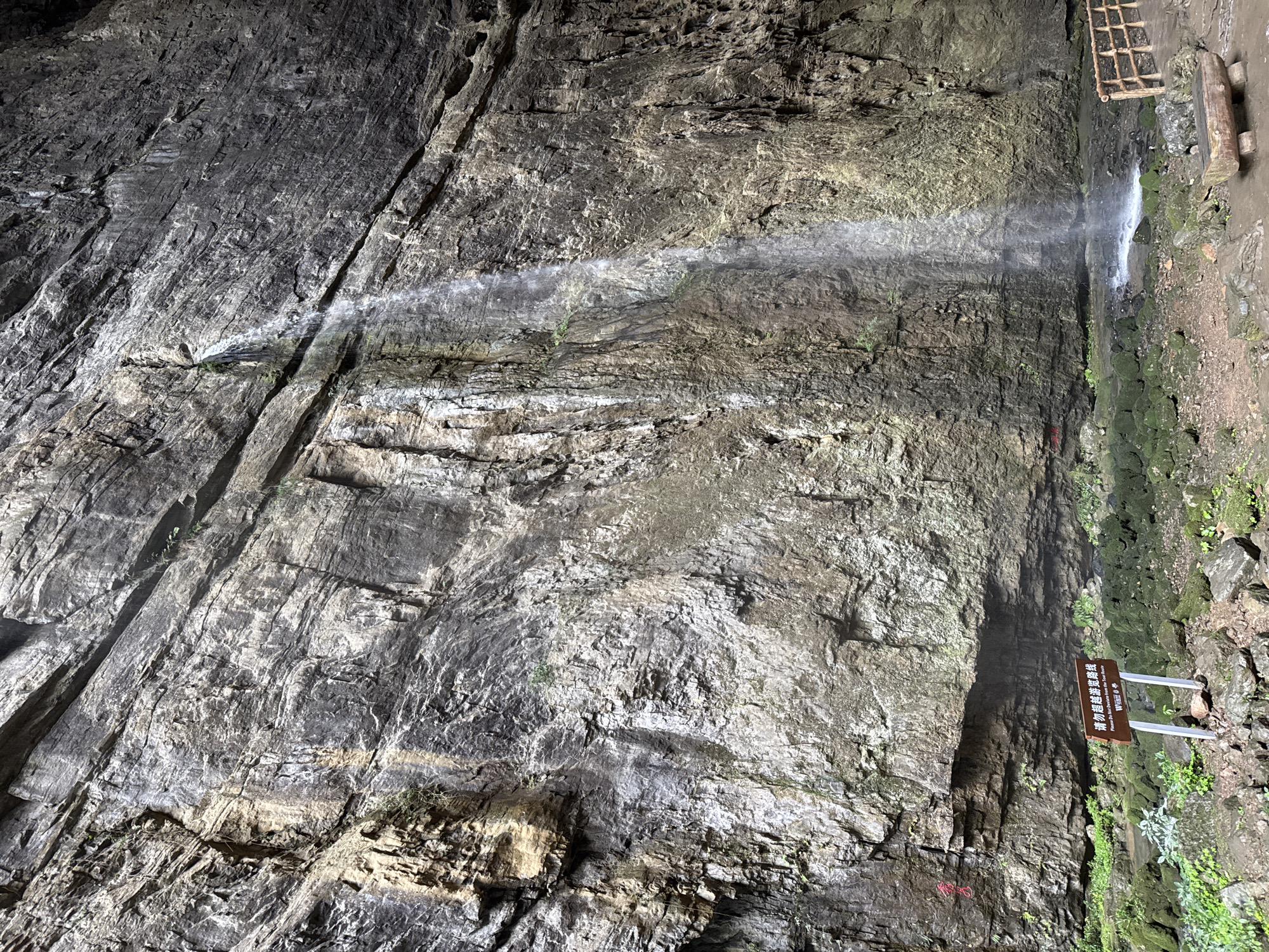 A horizontal image shows a rock wall and a small waterfall.
The wall is rocky with different shades of gray and brown, as well as a dark fissure running from the top left corner to the bottom left corner. A thin waterfall runs horizontally across the image to the right and then vertically down the right side. On the ground on the right is a sign with Chinese writing and plants growing. Above the sign is a structure that looks like a wooden fence or ladder.
The location is identified as Wulong District, China.