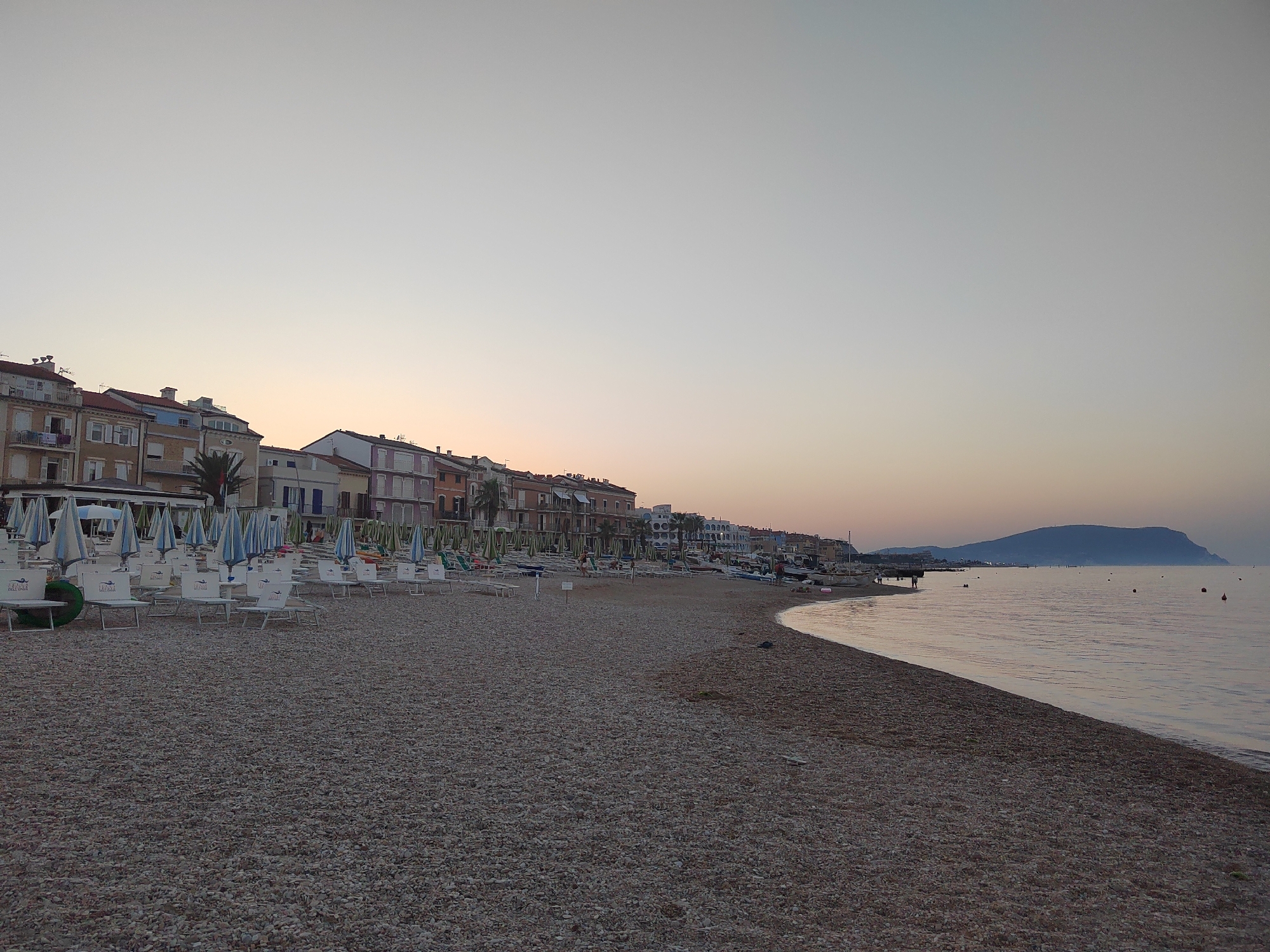The image depicts a serene sunrise over the pebble beach of Porto Recanati, Italy. Buildings line the shore, their facades painted in varying shades of beige, orange, and pastel colors. A row of closed umbrellas and empty lounge chairs are set up on the beach, suggesting the early hour before visitors arrive. The calm sea stretches towards the horizon, where a distant, hazy hill is visible. The sky is a soft gradient of pale blue and peach, indicative of the dawn. A few buoys dot the water, and a small pier extends into the sea on the right side of the frame. The overall impression is one of tranquility and the quiet beauty of a coastal town at daybreak.