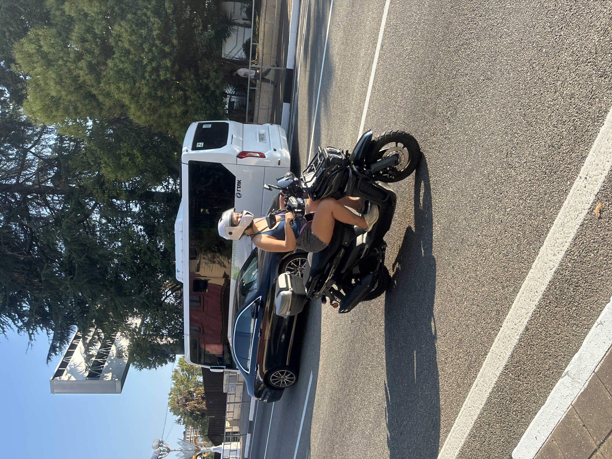 An outdoor, eye-level, medium shot captures a woman riding a black scooter on a sunny day in Sochi, Russia. She is wearing a white helmet, a dark blue tank top, and grey shorts. The scooter has a top case behind the seat. In the background, a black Mercedes sedan is parked next to a white bus. The bus has the letters "ГПЛК" on its side, indicating it is likely a public transport vehicle. A man in a light-colored shirt and dark pants walks on a sidewalk behind the bus. The scene takes place on an asphalt road with white lane markings. Trees and some architectural structures are visible in the background, suggesting an urban environment. The sky is clear and blue.