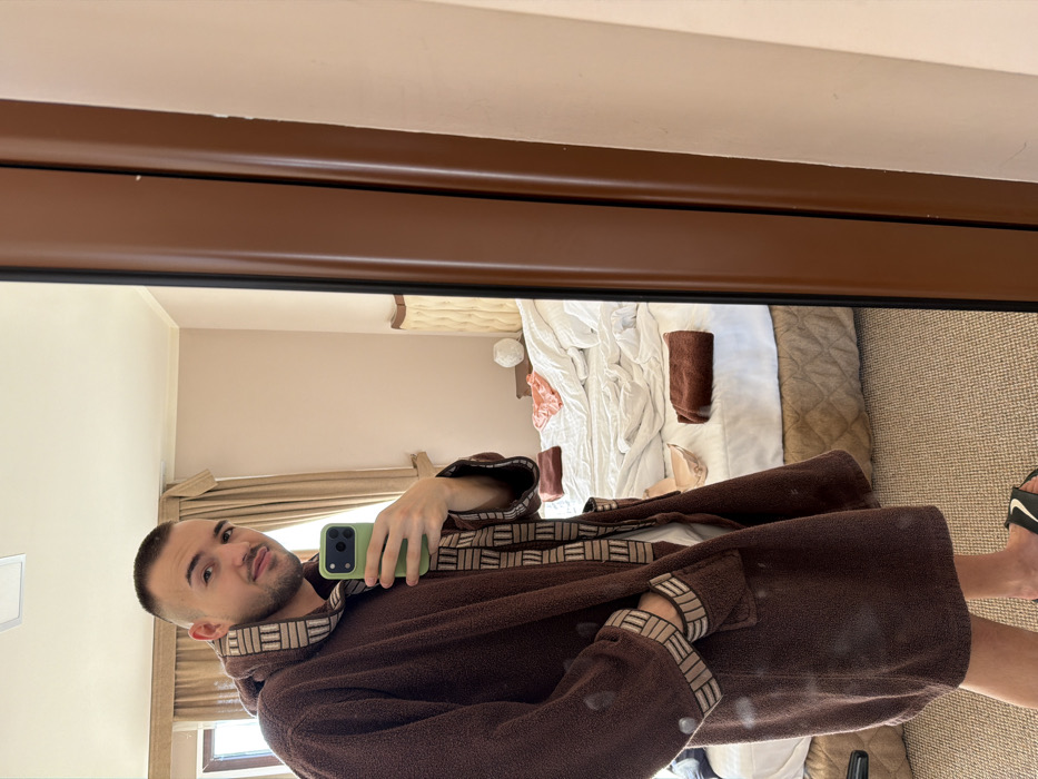 A young adult male is depicted taking a mirror selfie in what appears to be a bedroom or hotel room in Kladnitsa, Bulgaria. The male, with a short buzz cut and light facial hair, is seen from the waist up, wearing a brown terry cloth bathrobe with a beige and brown patterned trim on the collar, cuffs, and pockets. He holds a light green smartphone in his right hand, positioned to capture his reflection, and his left hand is in his bathrobe pocket. He is looking towards the camera with a slight smile. The background, visible in the mirror's reflection, reveals a disheveled bed with white bedding, a beige headboard, and a quilted beige bed skirt. A folded brown towel, a light pink garment (possibly underwear), and another lighter-colored garment (possibly a bra) are present on the bed. A small nightstand with a white spherical lamp is located next to the bed. A window with light brown curtains is visible on the left side of the reflection. The room features light pink-beige walls and a light beige carpeted floor. The mirror itself has a prominent brown wooden frame. The male also wears black and white flip-flop style sandals.