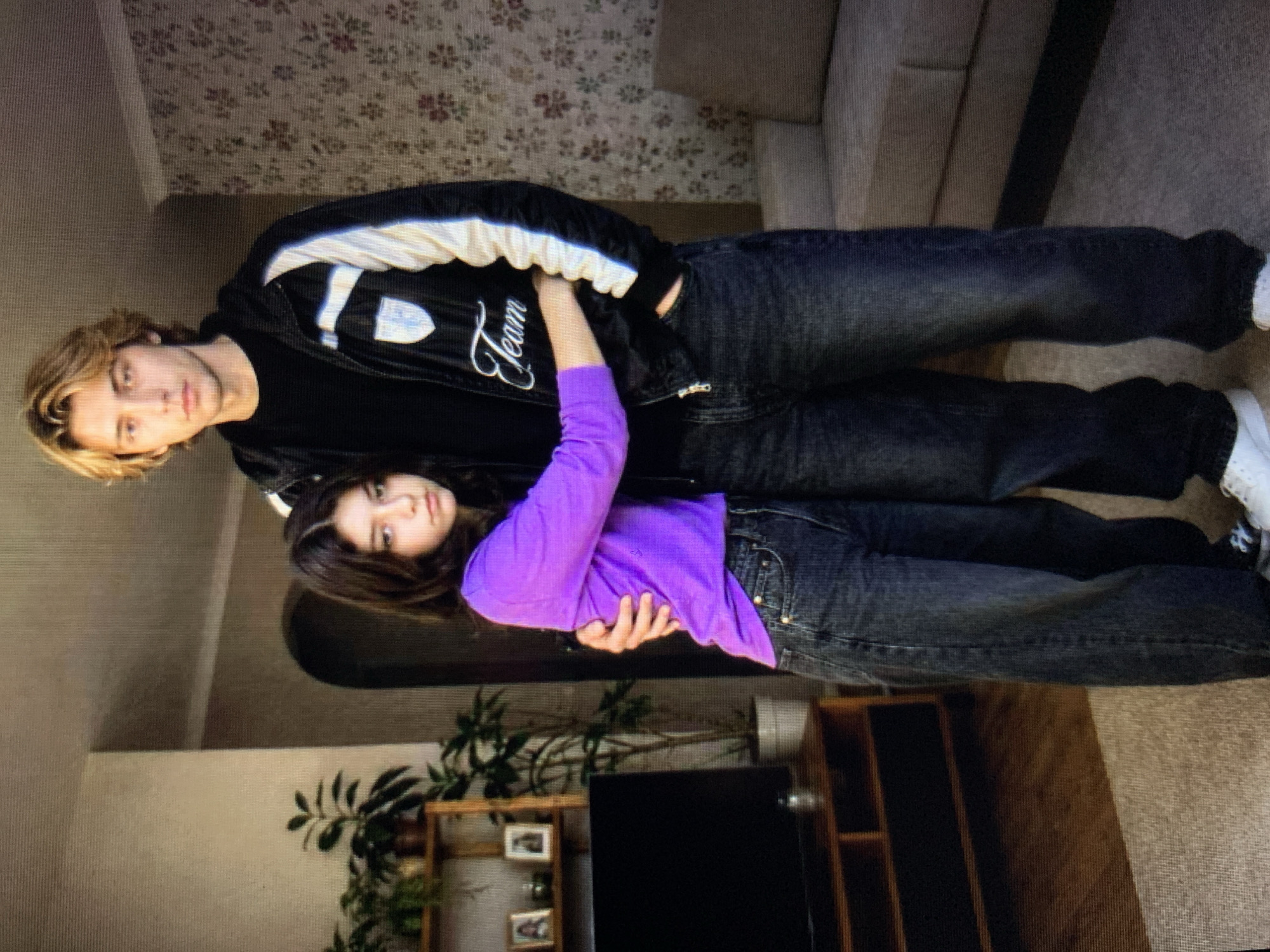 In a domestic interior, possibly in Lymanka, Ukraine, a young man with blonde hair stands embracing a young woman with dark hair. The man is wearing a black jacket with white stripes and the word "Team" embroidered on it, over a black t-shirt, and dark wash jeans. The woman is wearing a vibrant purple long-sleeved shirt and dark wash jeans. They are standing in what appears to be a living room, with a television stand and a shelf with plants and framed pictures visible in the background. A patterned wallpaper and a sofa are also present in the scene. The lighting suggests an indoor setting during the day.