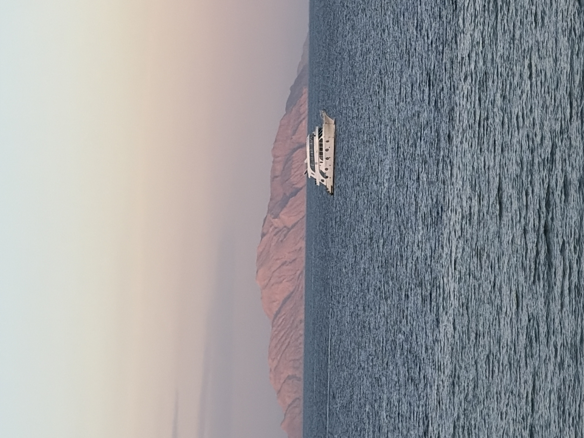 The image displays a maritime scene in Sharm El Sheikh, Egypt, likely depicting Tiran Island in the distance, observed from the coastline. The foreground consists of dark blue-grey water with fine surface ripples, occupying approximately the lower two-thirds of the frame. A white, multi-deck motor yacht is positioned in the mid-ground, slightly right of center, heading leftwards across the water; a faint wake trails its stern. The yacht features multiple circular portholes on its main hull and an enclosed upper deck structure. In the background, a large, arid mountain range or island stretches horizontally across the horizon, exhibiting soft contours and illuminated by warm, pinkish-orange light. The sky above, comprising the top third of the image, presents a gradient of pastel hues, transitioning from light blue at the zenith to pale peach, pink, and lavender closer to the horizon, consistent with either sunrise or sunset conditions. No individuals are visible on the boat or within the immediate environment.