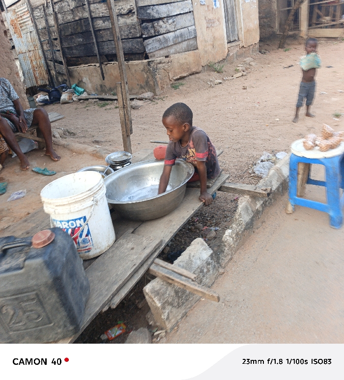 A child washes in a large metal bowl on a makeshift table over a ditch. Nearby, a blue plastic stool holds items under a sunlit sky, and another child stands in the background holding an object. In the background, an adult sits while several buildings and items are observable. The scene depicts an everyday moment within what seems to be a modest rural location.