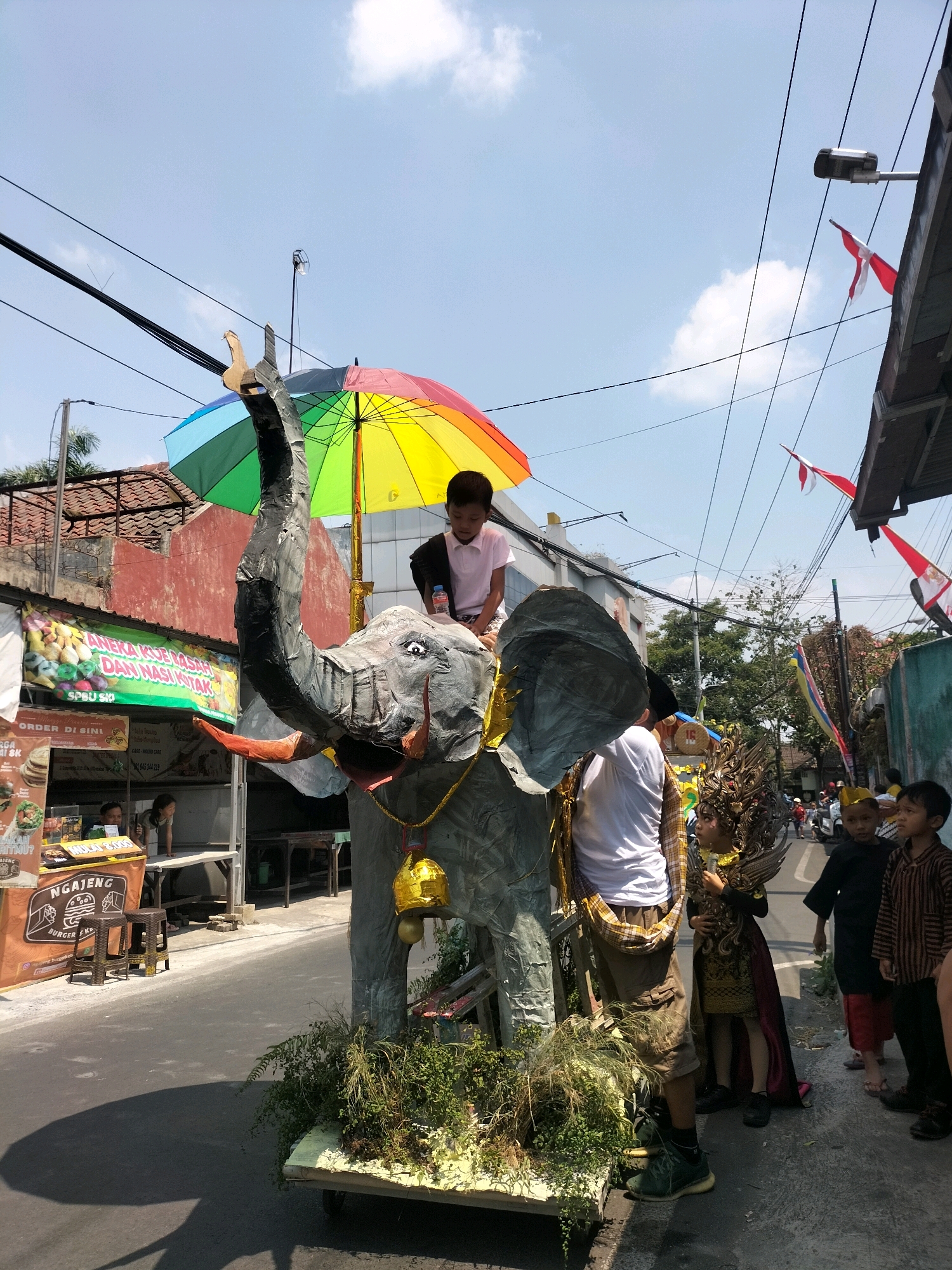 The image depicts a lively street scene in Malang, Indonesia, likely during a festive event or parade. In the foreground, a large, grey, elephant-shaped float dominates the frame, adorned with a colorful umbrella and a golden bell. A young boy in a white shirt is seated atop the elephant's head.
To the right of the float, several people are visible. A man in a white t-shirt and khaki shorts stands next to the elephant, and a young person in an elaborate costume with a headdress stands beside him. Further to the right, a group of children are gathered, one in a black shirt and red pants, and another in a striped brown shirt. Red and white flags are strung overhead, along with a streetlamp.
The background shows a street lined with buildings. On the left, a food stall with signage in Indonesian is visible, displaying images of food items. The street itself is paved, and the sky is bright blue with scattered clouds, suggesting a sunny day. The overall impression is one of community celebration and cultural activity.