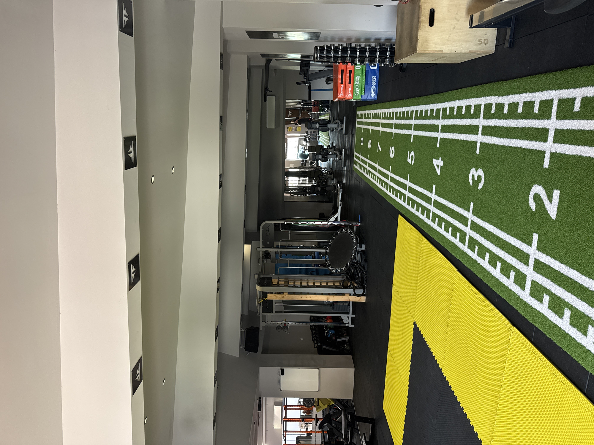 The image shows the interior of a gym in Tsarevo, Bulgaria. A central, green, artificial turf runway marked with white lines and numbers (2 through 8) runs along the right side of the image. Yellow interlocking floor tiles are to the left of the turf. Various pieces of exercise equipment line the left side of the gym, including weight racks, a functional trainer, and a whiteboard. Dumbbells are stacked on a rack at the far end. The ceiling has a series of recessed lights. Arrows point to the right on wall posts.