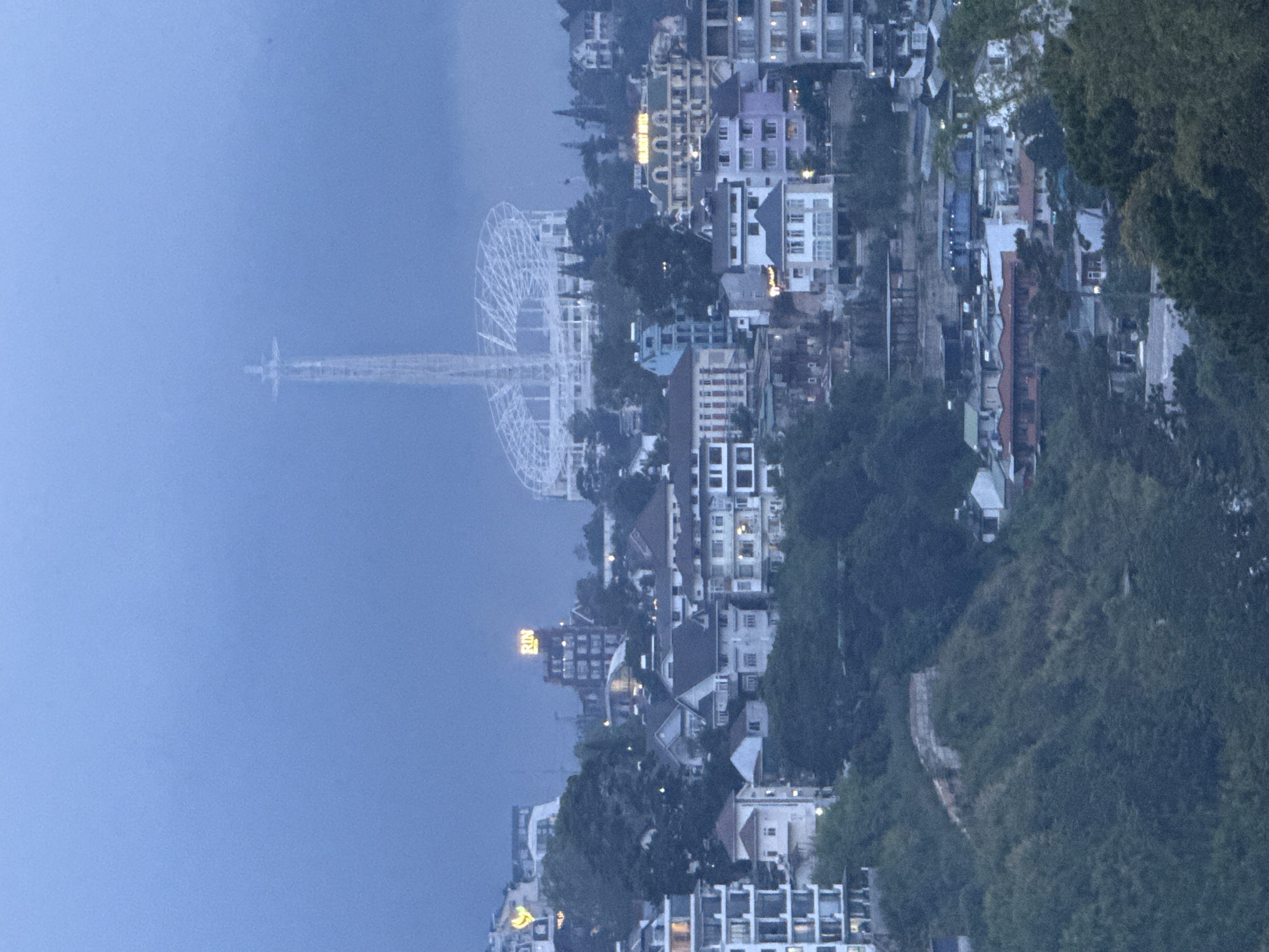 The image shows a panoramic view of the city of Da Lat, Vietnam, at dusk. The sky is a hazy blue, and a large, white statue of Jesus Christ is visible in the distance, towering over the city. The statue is illuminated, and its silhouette stands out against the darkening sky. Below the statue, a white, circular structure, likely a stadium or amphitheater, is also visible.
The city itself is built on a hillside, with buildings cascading down the slopes. The architecture is a mix of styles, with some modern buildings interspersed with older, more traditional structures. Many of the buildings have brightly lit windows, suggesting that people are home despite the approaching night. The overall impression is one of a vibrant, bustling city nestled within a natural landscape.
In the foreground, lush green trees and vegetation cover the hills, adding to the natural beauty of the scene. The image captures the unique charm of Da Lat, a city known for its French colonial architecture, misty mountain air, and stunning natural landscapes.