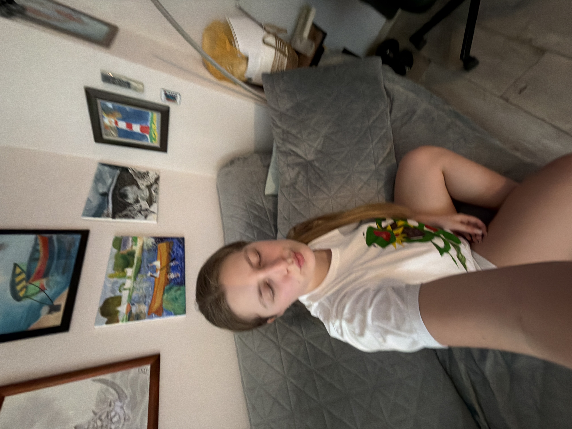 The image captures an intimate indoor scene, likely a living room or bedroom, where a young female is resting on a grey quilted sofa or bed. She has long, light brown hair flowing over her shoulder and is wearing a white t-shirt with a colorful, possibly abstract or animal-themed, design on the front. Her eyes are closed, indicating she is either asleep or in a state of deep relaxation.
The room features light-colored walls decorated with several artworks. Visible on the wall are a framed picture of a red and white striped lighthouse against a blue sky, and another framed painting depicting a boat with a red and yellow sail on blue water. Below these are two smaller, unframed canvases: one features a dark, possibly abstract or natural scene, and the other shows a vibrant landscape with green trees and a building or structure. A larger, partially visible framed picture with a brown frame at the bottom appears to depict the head of an animal, possibly a ram or goat.
In the upper part of the frame, a light-colored ceiling fan is partially visible, and on a shelf above the sofa, there's a decorative wooden basket or item. The floor, visible towards the right, appears to be made of light-colored wood or laminate, with the black legs of a chair also visible. The lighting suggests it is daytime. The overall atmosphere is serene and personal. There is no discernible text visible in the image, nor are there any specific visual cues to confirm the location as Burgas, Bulgaria.