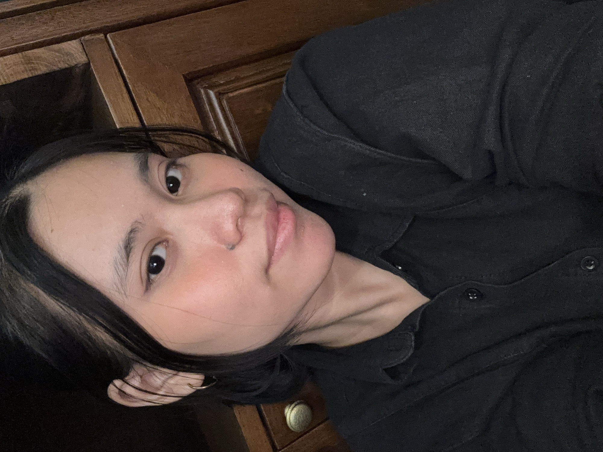 The image is a close-up, selfie-style photograph of a young person with dark hair and fair skin. They are wearing a black collared shirt. The background appears to be a wooden cabinet with a brass knob. The lighting is warm and casts a slight shadow. There are no recognizable landmarks that would indicate a specific location within South Jakarta, Indonesia. The scene is static and focused on the individual's face and upper torso.