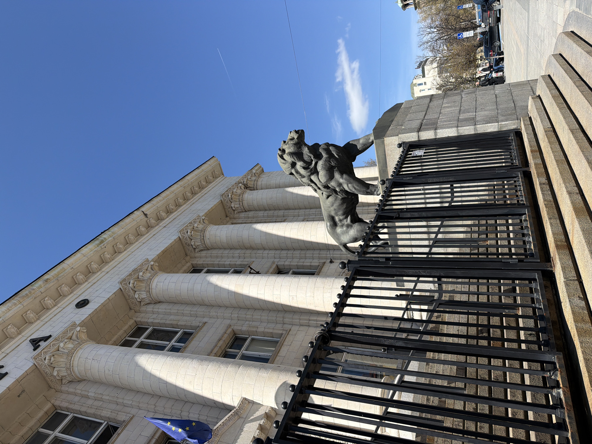 The image depicts a lion statue flanking the entrance to the National Assembly building in Sofia, Bulgaria. The statue is made of dark metal, and is mounted on a grey stone plinth. The metal gates of the building are in the foreground, and the stone steps that lead up to the gates are visible on the right side. The building itself is white and features a classical architectural style, with columns. An EU flag is visible on the lower left of the building. The sky is blue, and there is a contrail visible. There are other buildings and vehicles in the background. The scene is bathed in sunlight, suggesting it is daytime.