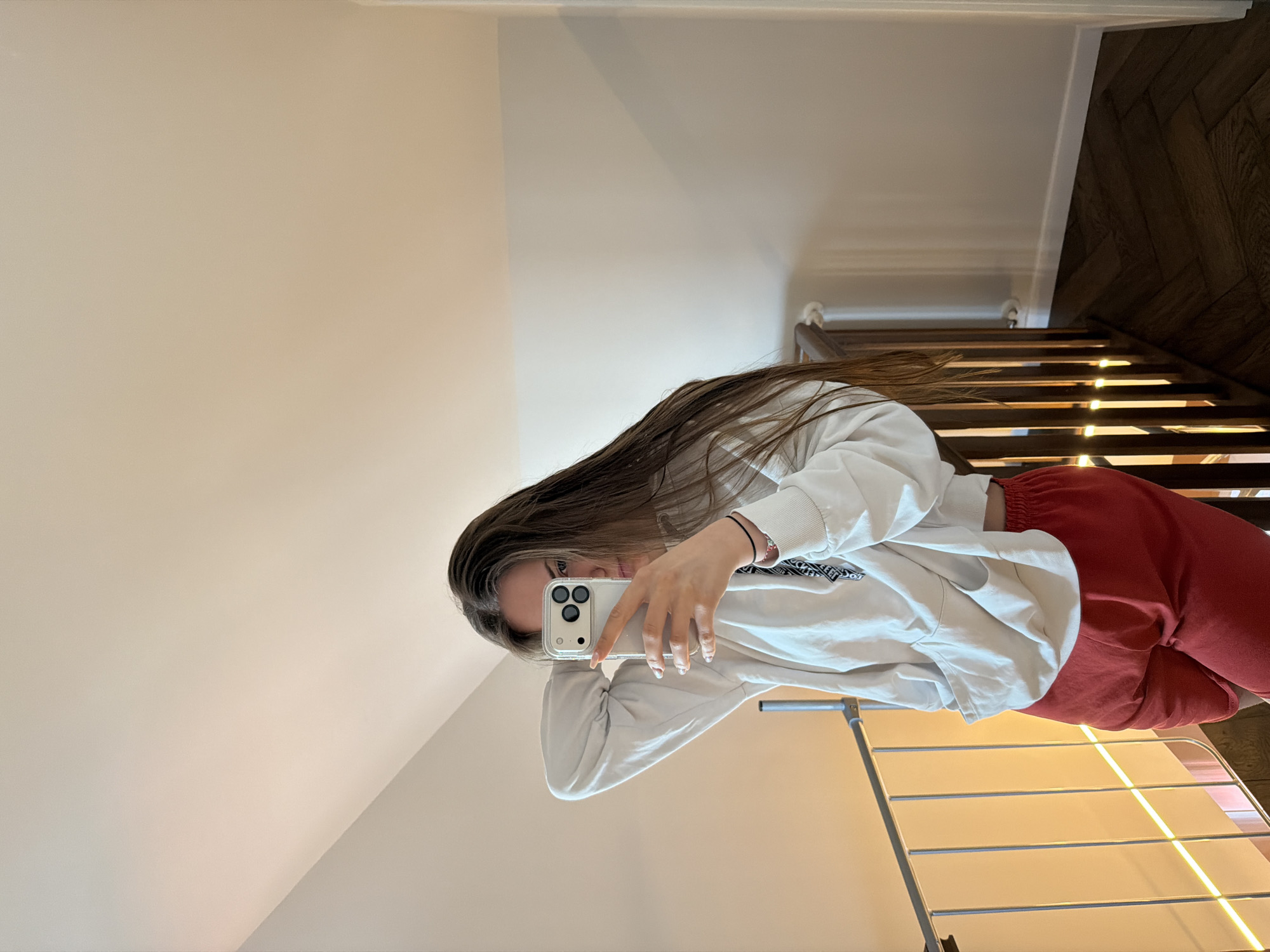 A young woman with long, brown hair is taking a selfie indoors, near a staircase. She is holding a white smartphone in front of her face, covering most of it. She is wearing a white hooded sweatshirt and red sweatpants. Behind her, there is a white wall, a wooden staircase with illuminated steps, and a metal drying rack. The location appears to be a residential interior, but there is no information in the image to confirm that this photo was taken in Buhovo, Bulgaria.
