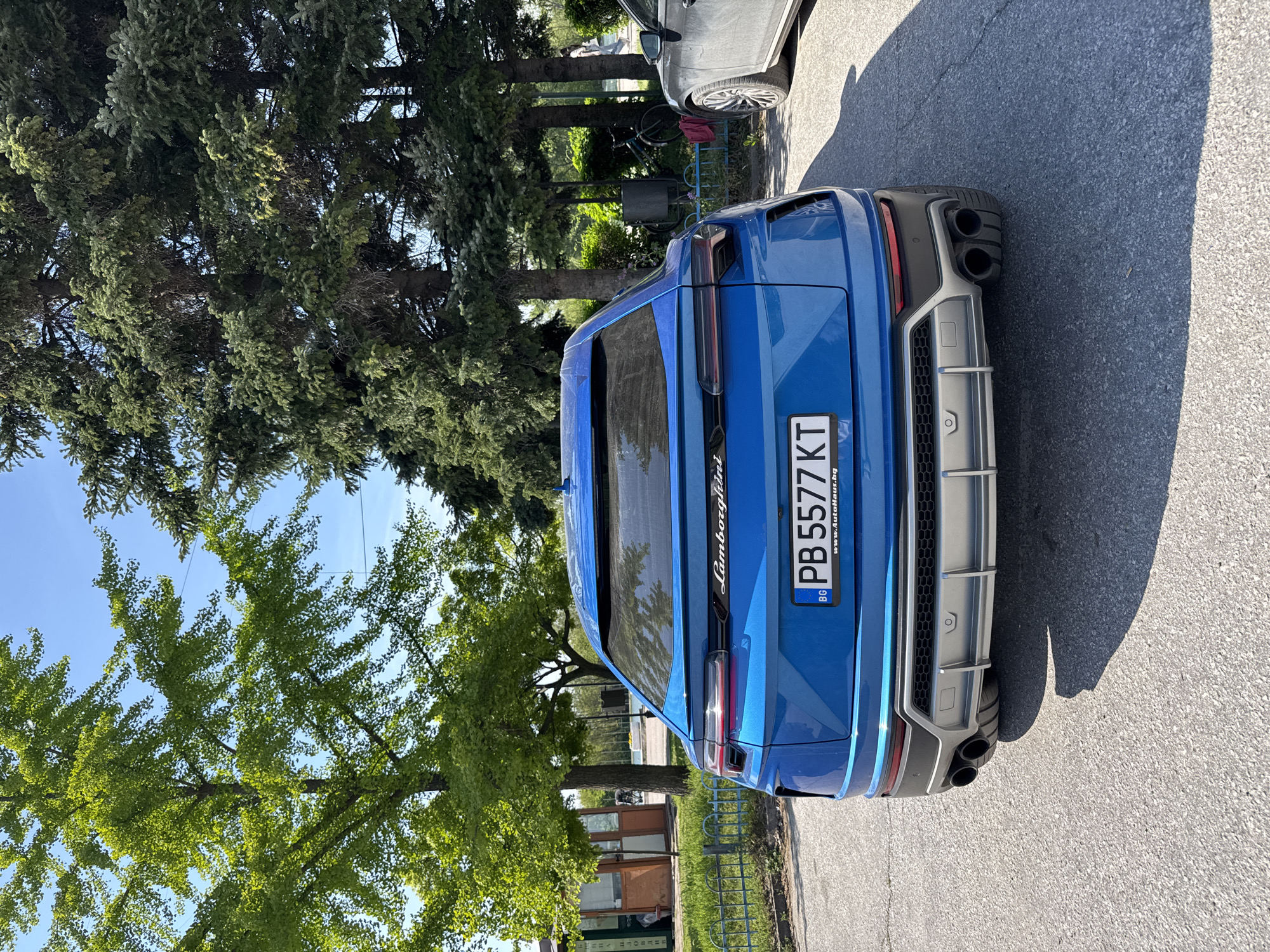 The image captures the rear view of a bright blue Lamborghini Urus, parked outdoors on a paved surface in what appears to be a street or parking area in Plovdiv, Bulgaria. The car is oriented with its rear facing the viewer, slightly angled. It features a distinctive design with a sporty rear diffuser in grey and black, housing quad exhaust pipes. The rear window prominently displays the "Lamborghini" script.
The Bulgarian license plate, "PB 5577 KT," is clearly visible, with "PB" indicating the Plovdiv region. Below the license plate, part of a car dealership website, "www.Autohaus.bg," is discernable on the plate holder.
The setting suggests a sunny day, with strong shadows cast on the asphalt, indicating a clear sky and bright conditions. Various types of trees with lush green foliage, including some coniferous and some with lighter green leaves (possibly ginkgo, as hinted by the crops), surround the area, creating a natural canopy and background. A clear blue sky is visible through gaps in the trees.
To the left of the Lamborghini, a portion of a grey car is visible, parked further ahead. In the mid-ground, behind the cars and a metal fence, there are more trees, a partially visible bicycle, and a small, rustic wooden structure or booth. The overall scene conveys a pleasant, bright daytime atmosphere.