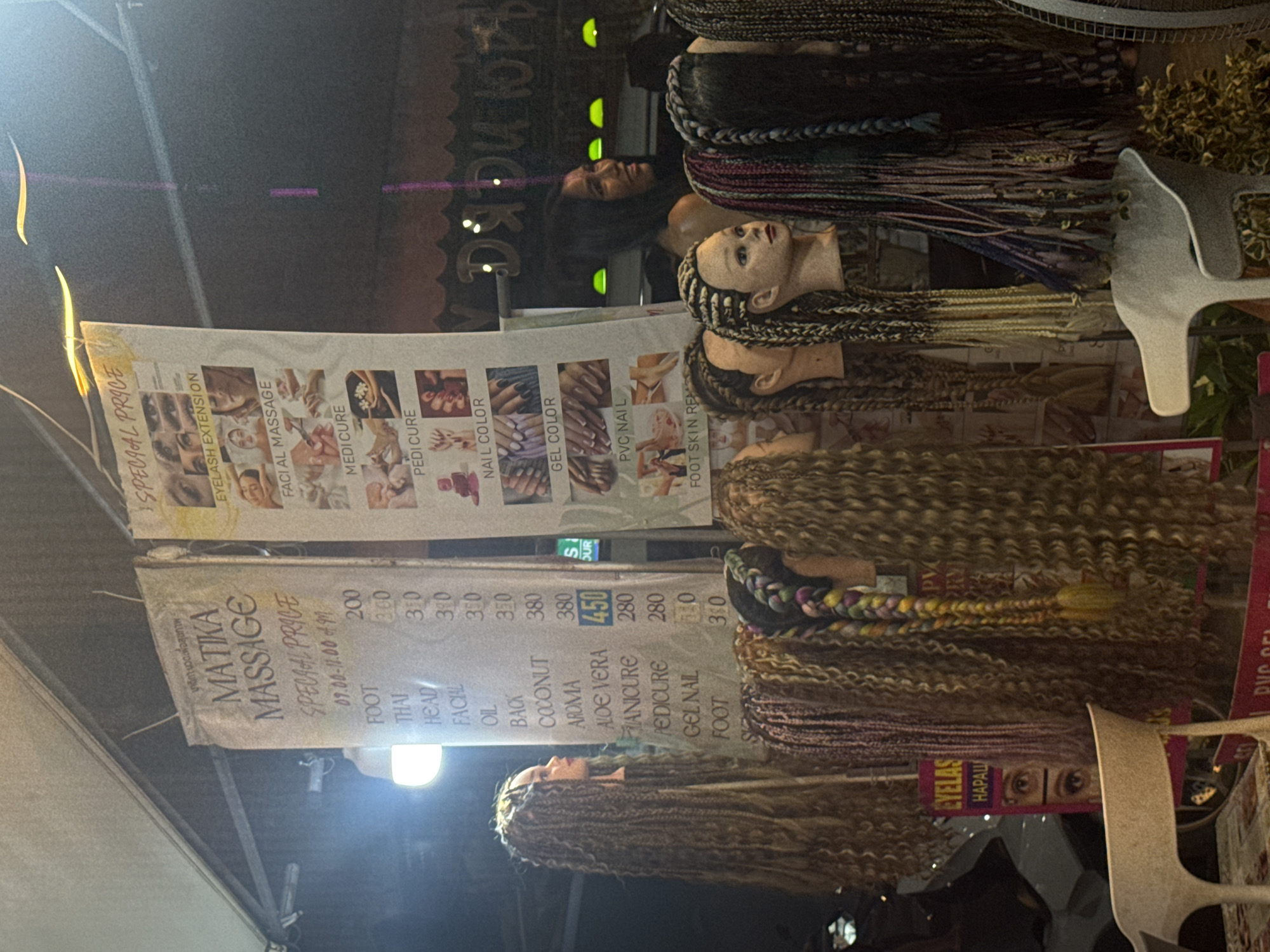 The image shows a street scene in Patong, Thailand, at night. There are mannequins with various types of braided wigs. Next to the mannequins, there are two signs advertising "Matika Massage" with a list of services and prices, including special prices available from 9:00 AM to 11:00 AM. The services listed include foot massage, Thai massage, head massage, facial massage, oil massage, back massage, coconut massage, aloe vera massage, manicure, pedicure, gel nail polish, foot skin repair, and eyelash extensions. There are people in the background of the image.