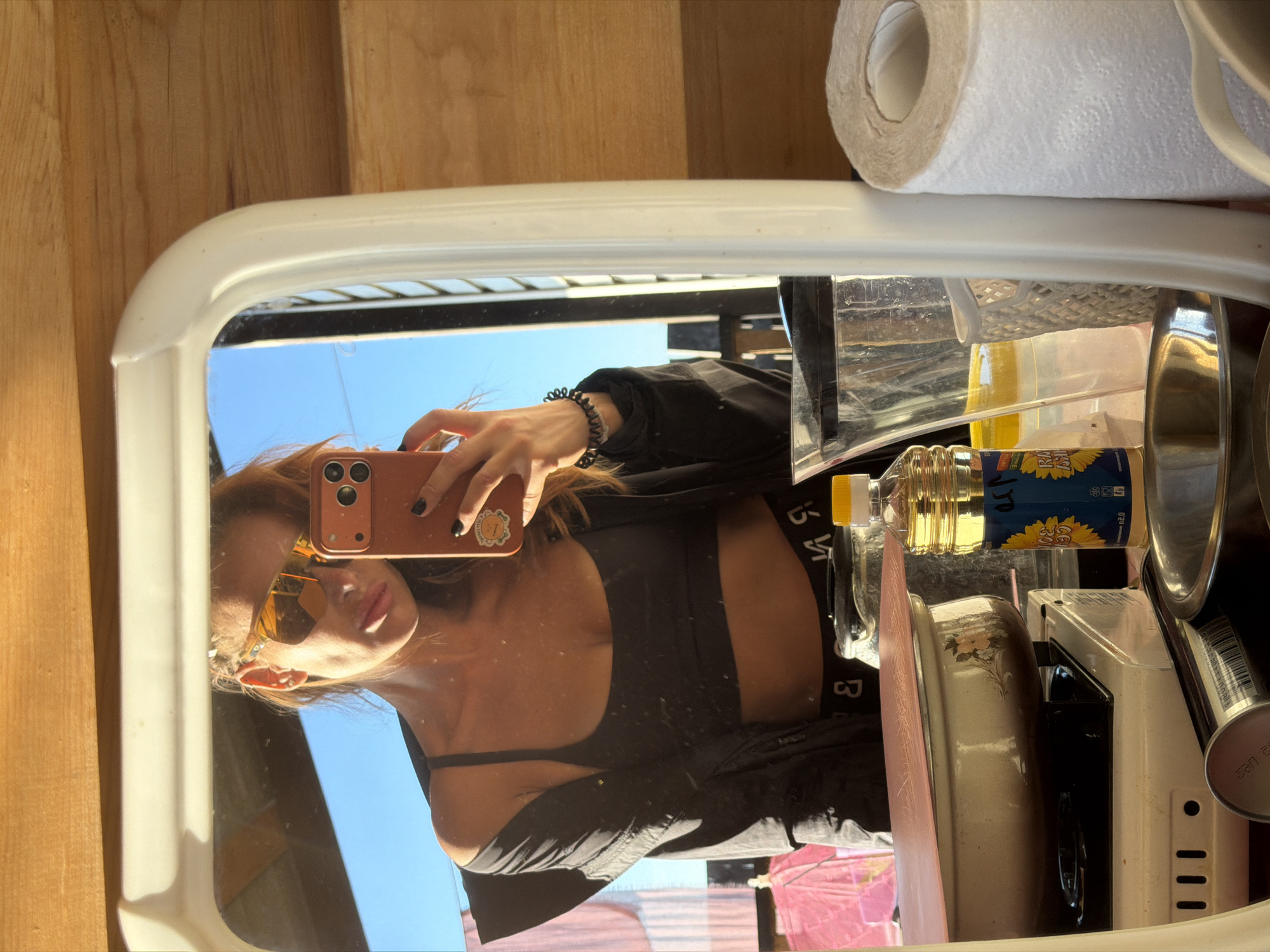 A vertical mirror reflects a young woman taking a selfie. She is wearing sunglasses and a black top. She is holding her phone in her left hand, which is in the upper left corner of the frame. The mirror is in a white frame. Behind the woman in the reflection, there are various objects, including a roll of toilet paper, a bottle of cooking oil, and some kitchenware. The background of the reflection is a bright blue sky, indicating it is daytime. The woman appears to be in an indoor setting, possibly a kitchen or bathroom, given the items reflected. There is no discernible text visible in the image. The overall mood appears casual and relaxed.