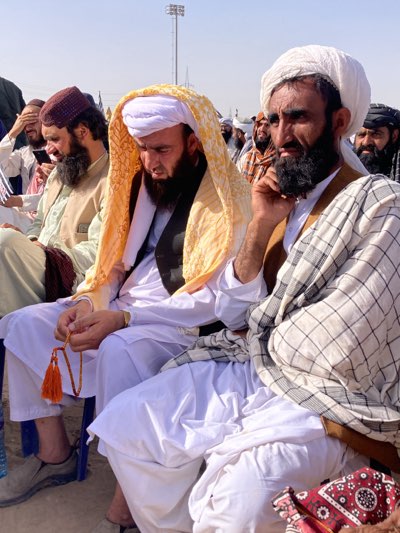 This image depicts an outdoor gathering of men in Pishin, Pakistan, under a bright, clear daytime sky. In the foreground, three men are prominently featured, seated on chairs.
The central figure wears a white shalwar kameez, a white turban, and a distinctive golden-yellow patterned fabric draped over his head and shoulders. He holds a string of prayer beads (tasbih) with orange tassels in his hands, looking down with a contemplative or prayerful expression. To his right, another man, sporting a full beard, wears a white turban, a brown waistcoat over a white garment, and has a white and black checkered shawl draped across him. He gazes intently towards the right, appearing focused. To the left of the central figure, a third man with a beard wears a maroon cap and an olive-green top, looking down at his hands.
Numerous other men, similarly dressed in traditional attire, are visible in the blurred background, indicating a larger assembly. The ground beneath the men appears to be unpaved, suggesting an open field or a dusty public area. A tall light pole or tower is visible in the distance, hinting at an organized outdoor space. The overall atmosphere suggests a solemn or attentive gathering, possibly religious or community-related. There is no visible text in the image.