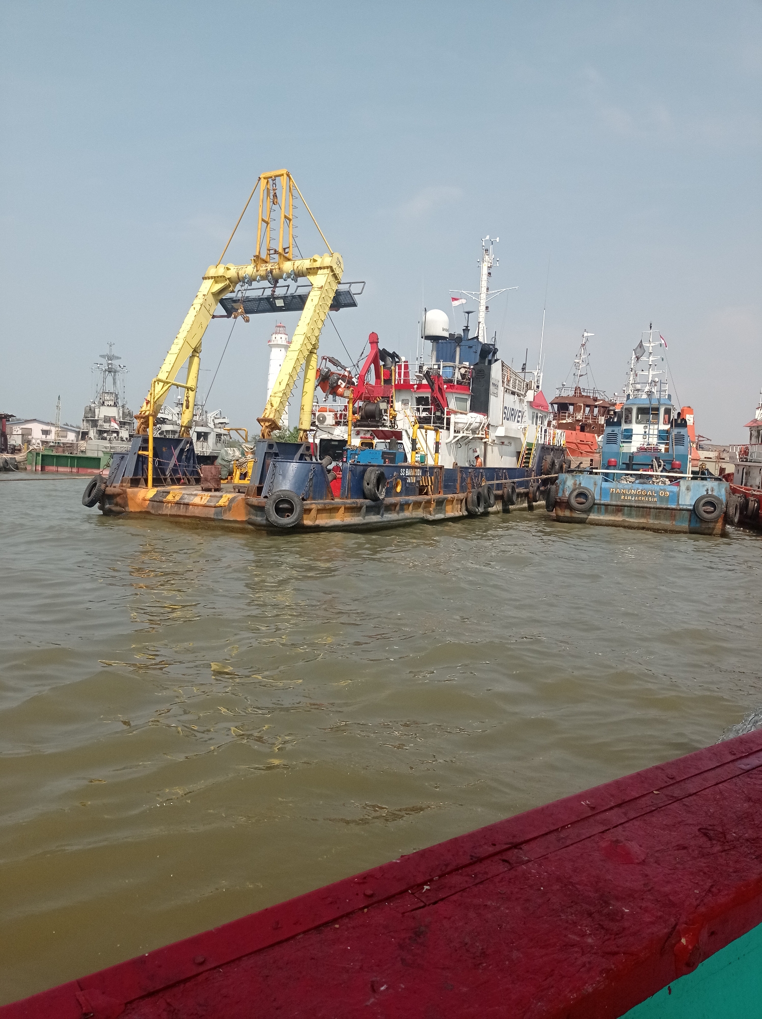 The image depicts a harbor scene in Tegal, Indonesia, characterized by several vessels docked in murky, brown water. In the foreground, the weathered red railing of a boat is visible. The central focus is a large, yellow gantry crane mounted on a barge, with its structure dominating the mid-ground. Adjacent to this barge, other vessels, including tugboats with the names "SS BATAKORA" and "MANUNGGAL 09" visible, are moored. A white lighthouse with red accents stands in the background, partially obscured by the gantry crane. The sky above is clear and light blue, suggesting daytime. Various other boats and structures, possibly part of the port infrastructure, are visible further in the distance. The scene appears to be an active port or industrial waterfront.