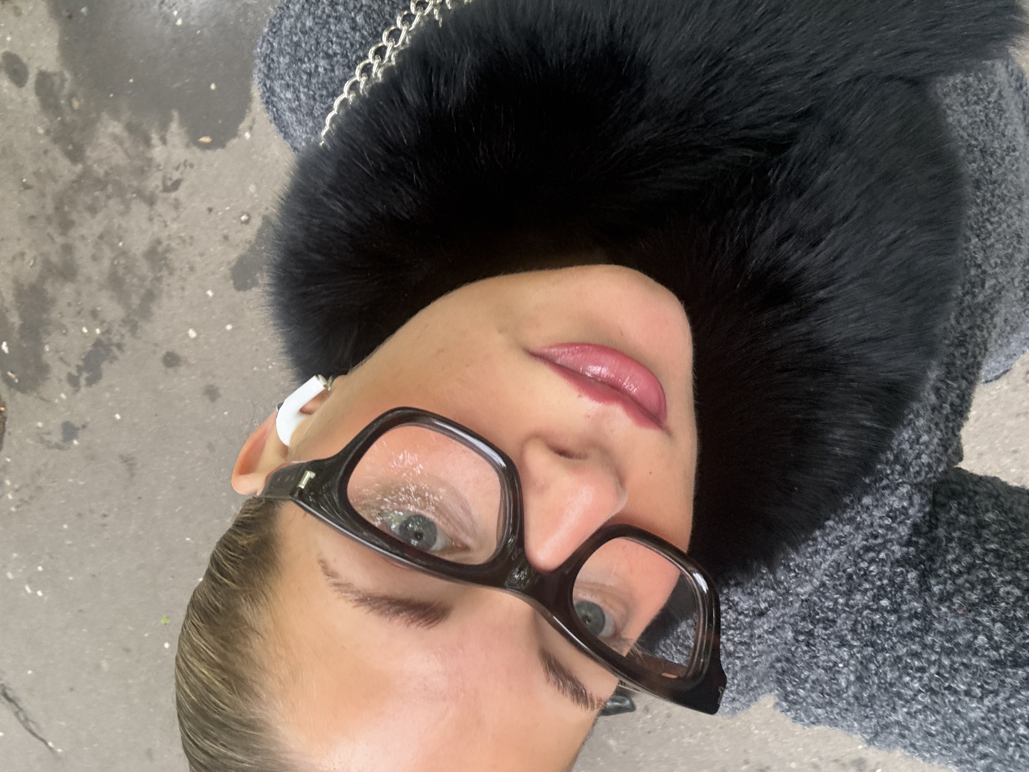 A woman with light skin, light-colored hair slicked back, and wearing large, dark-rimmed glasses, is centered in a downward-facing photograph. She has a white wireless earbud in her left ear. Her lips are pursed and appear glossy. She is wearing a black fur collar and a gray textured coat or jacket. The background is a gray concrete surface, possibly a sidewalk. The scene is likely in Paris, France, given the fashion style and common urban setting.