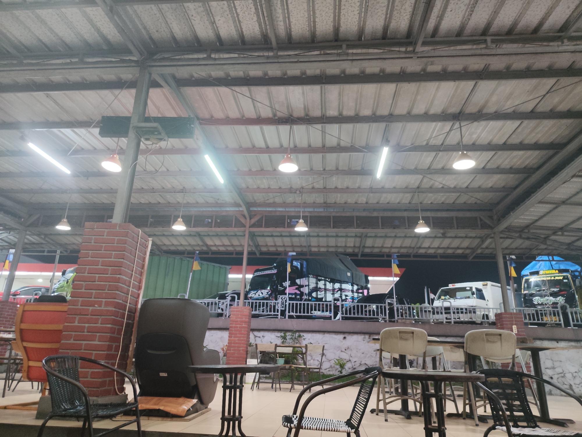 The image depicts an outdoor seating area under a corrugated metal roof supported by metal beams and poles. The area is illuminated by fluorescent lights and a few hanging lamps. In the foreground, there are several tables and chairs, including a black woven chair and a brown wooden chair. A red brick column stands to the left of the frame. In the background, several vehicles are visible, including a black truck with colorful graphics, a white van, and a green container. The scene appears to be a rest stop or roadside eatery in Nagrog, Indonesia. There are no people visible in the image.