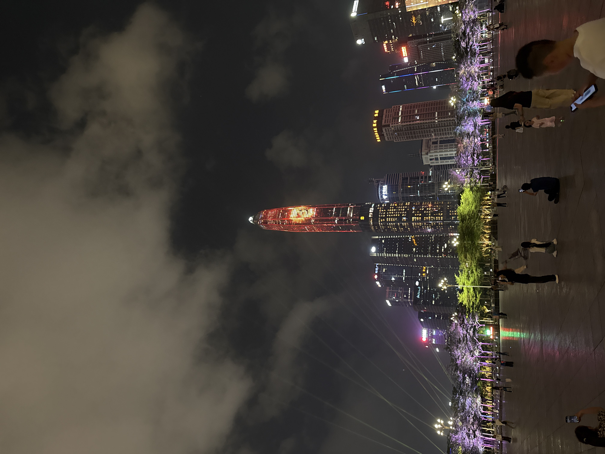 The image depicts a nighttime scene in Futian District, China, showcasing a cityscape dominated by illuminated skyscrapers. A particularly prominent red-lit skyscraper stands out against the cloudy night sky. In the foreground, a paved plaza is visible, where several people are present. Some individuals are engaged in taking photographs, while others appear to be strolling along the plaza. Trees, adorned with decorative purple lights, line the plaza's edge, contributing to the urban ambiance.
