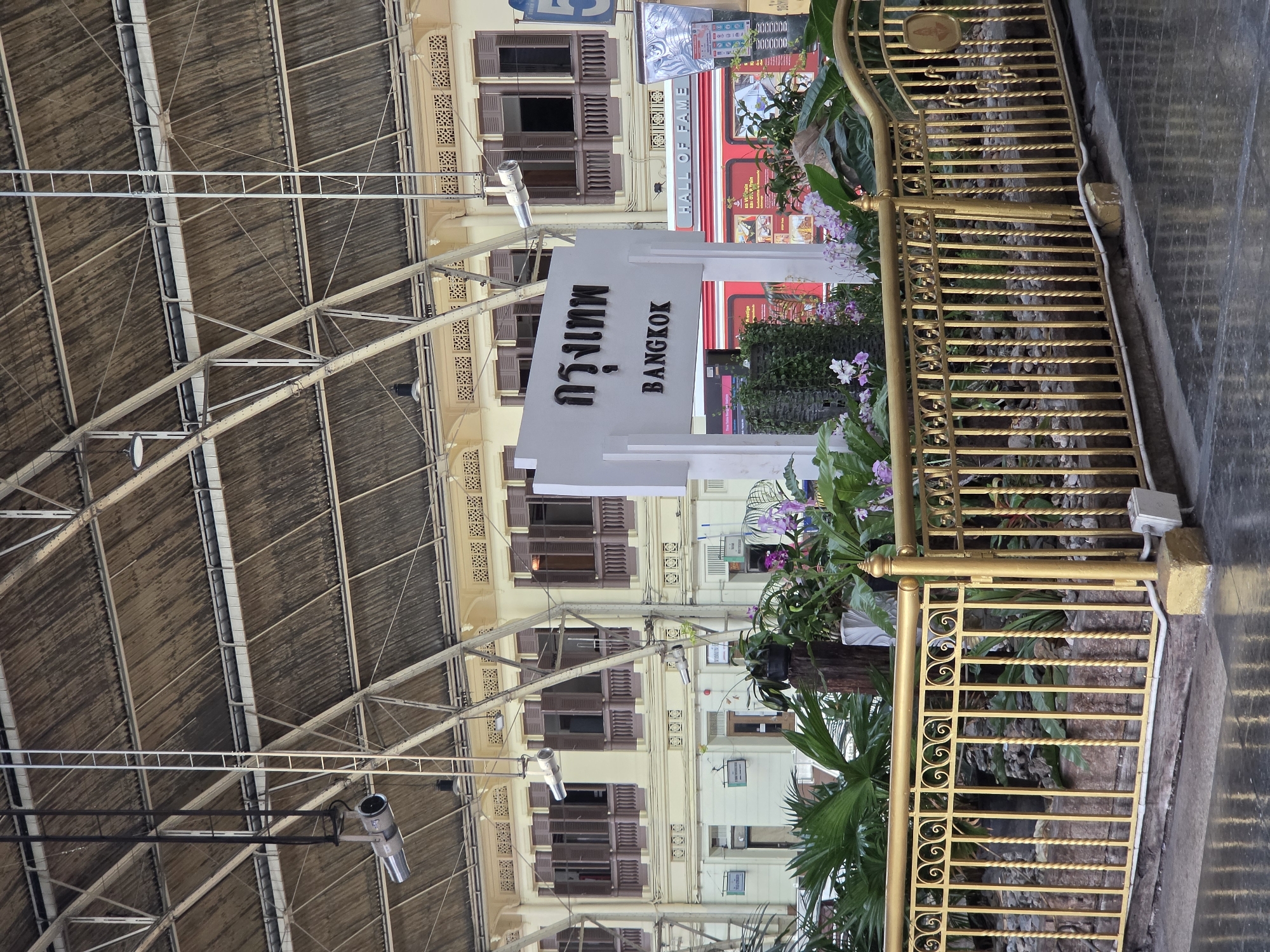 The image depicts the interior of Bangkok Railway Station (Hua Lamphong), Thailand, with a sign reading "BANGKOK" in both English and Thai script. A golden railing borders a planter filled with lush greenery and flowers. The station's high roof structure, supported by metal beams and corrugated material, is visible above. Several security cameras are suspended from the roof structure. Buildings with shuttered windows flank the station entrance.