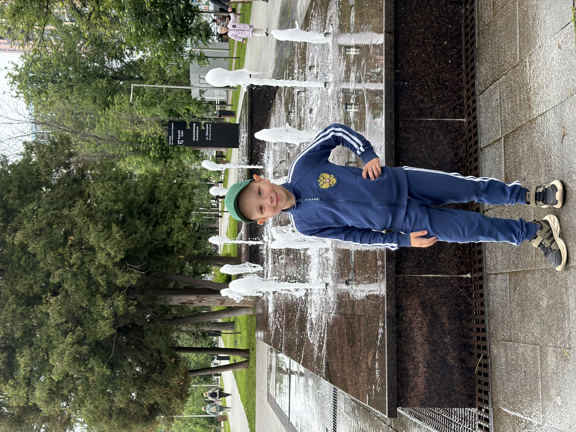 A young boy wearing a blue tracksuit and a green baseball cap stands in front of a public fountain in a city park. The fountain has several water jets spraying water upwards, creating a misty effect. The boy is smiling and has his left hand on his hip. The park appears to be well-maintained, with trees and paved pathways visible in the background. The setting suggests a public recreational area, possibly in a city, though the specific location in Perm, Russia, is not visually discernible from the image itself. The overall atmosphere is cheerful and relaxed, indicative of a pleasant day outdoors.