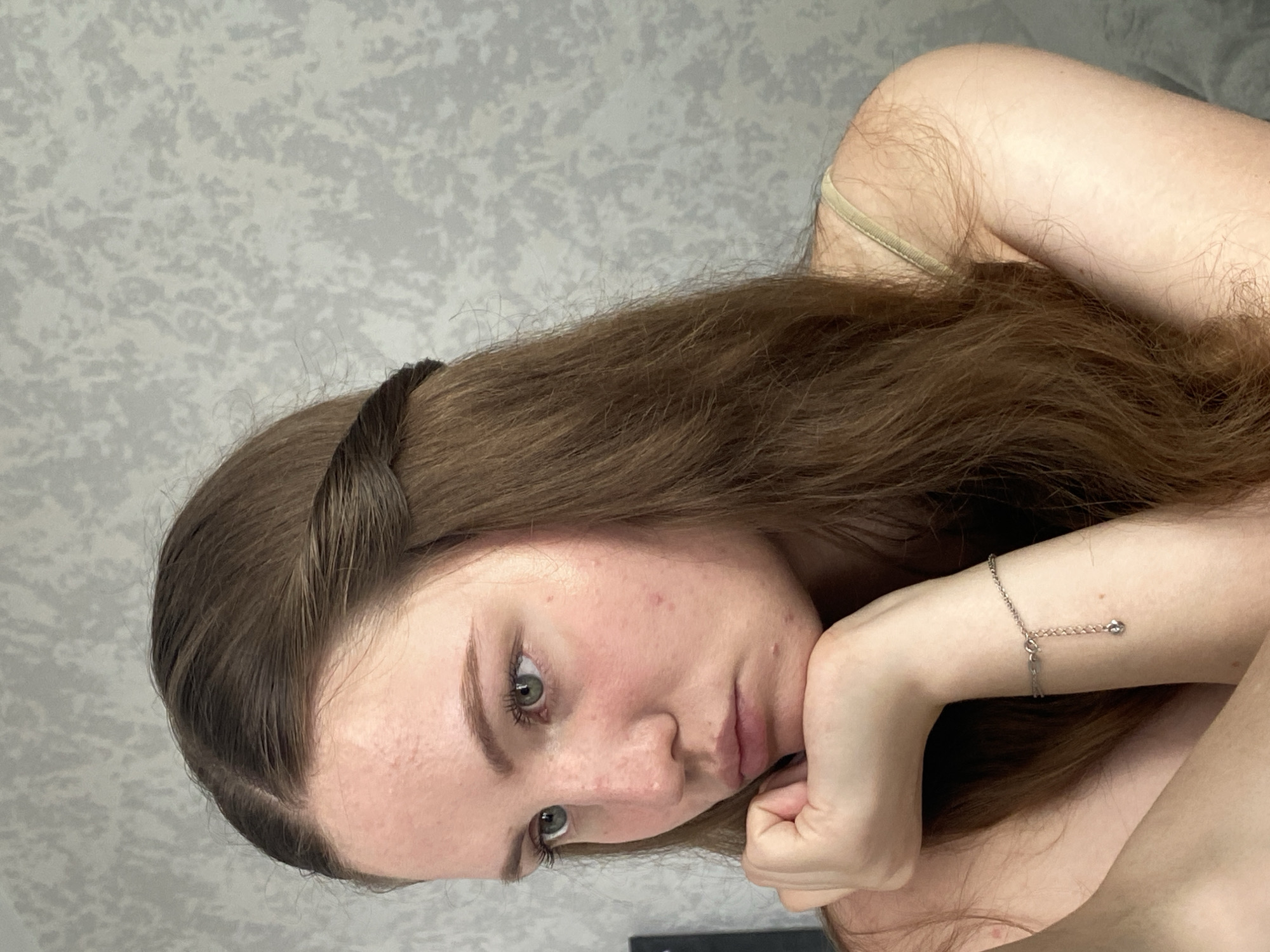 A young woman with brown hair and green eyes is shown in a medium shot, looking away from the camera with her chin resting on her hand. She is wearing a light-colored top and a silver bracelet with a small charm. Her hair is long and wavy, with a braided section across the top of her head. The background appears to be a textured wall. There is no visible indication of the specific location within Izhevsk, Russia.