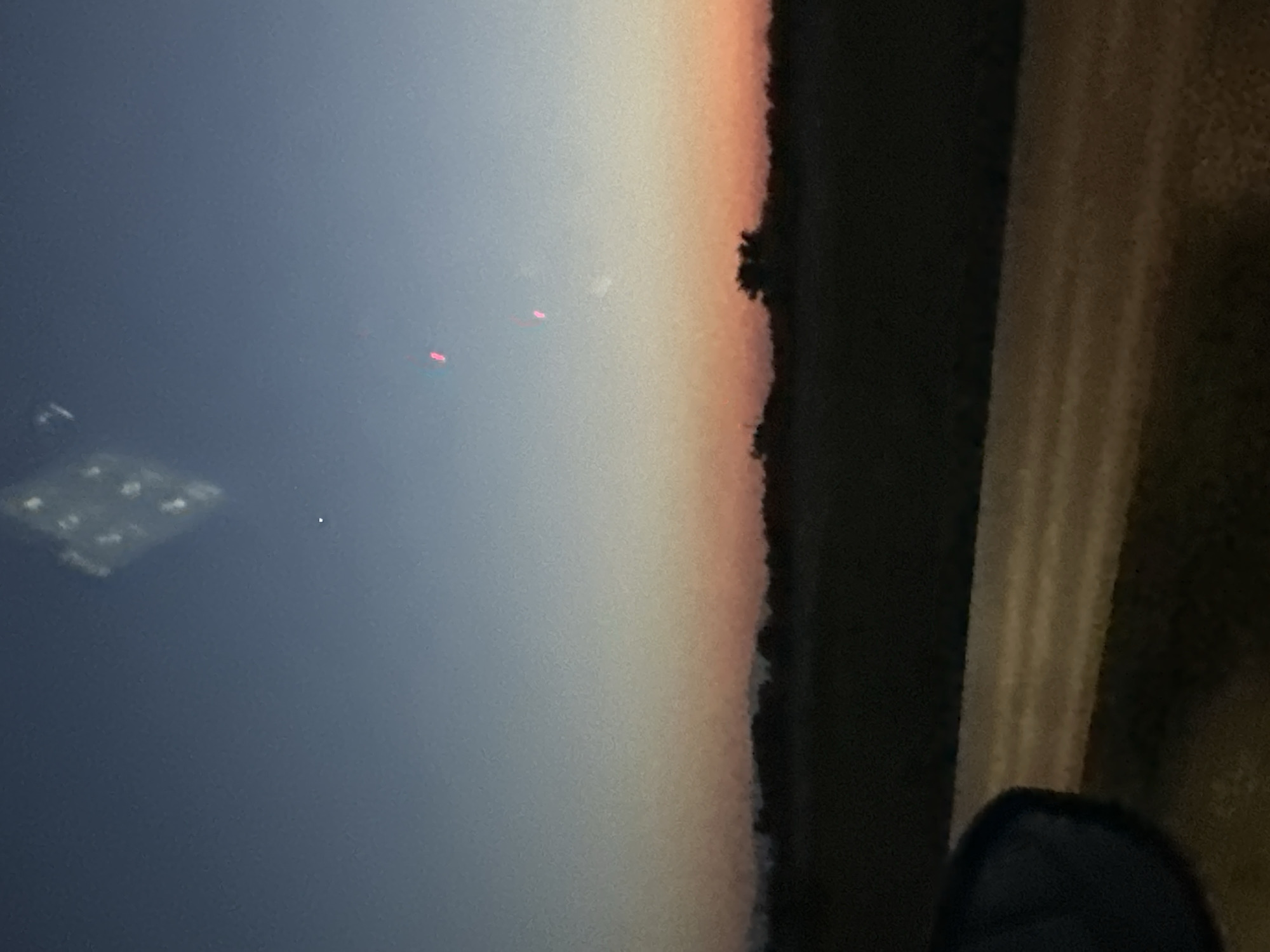 The image depicts a twilight sky over a rural landscape. The sky transitions from a deep blue at the top to lighter shades of orange and pink near the horizon. Silhouetted against the sky are trees and a low-lying landform. In the foreground, a dark, amorphous shape is visible on the left, possibly a car mirror or window. A road or path runs horizontally across the bottom half of the image, with faint stripes of light on its surface. There are a few small, bright specks in the sky, one appearing as a white dot and two others as vertical red lines. A blurry, diamond-shaped reflection is also present in the upper part of the sky. The scene appears to be taken from a moving vehicle. The location of Skutare, Bulgaria is not discernible from the visual information in the image.