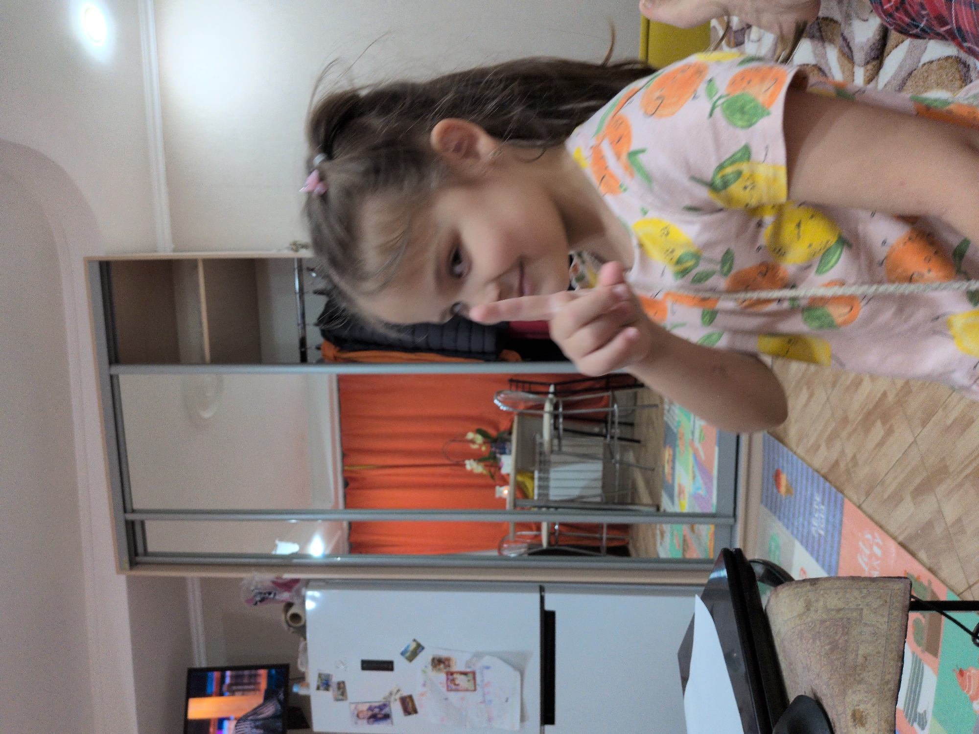 The image shows a young girl captured in an indoor domestic setting, identified as Sochi, Russia. The girl, with brown hair partially tied back with a pink clip, is looking towards the viewer with a slight smile and her right index finger extended, pointing. She is wearing a light pink short-sleeved t-shirt patterned with cartoon-like yellow lemons and orange citrus fruits.
The background features a large wardrobe or closet with mirrored sliding doors that reflect another part of the room. In the reflection, vibrant orange curtains are visible, along with a table and two chairs, suggesting a dining area or a similar seating arrangement. A small decorative plant or flowers sit on the reflected table. Below the mirrored doors, a white refrigerator adorned with various magnets, photos, and children's drawings is partially visible. Further to the left, a television screen displaying colorful visuals can be seen. The floor is tiled or covered with wooden-look planks, and a patterned mat with some indistinct text lies on it. A bright light fixture is visible on the ceiling in the upper left of the frame. The scene conveys a casual, lively atmosphere within a home. The time of day is not evident, but the interior is brightly lit.