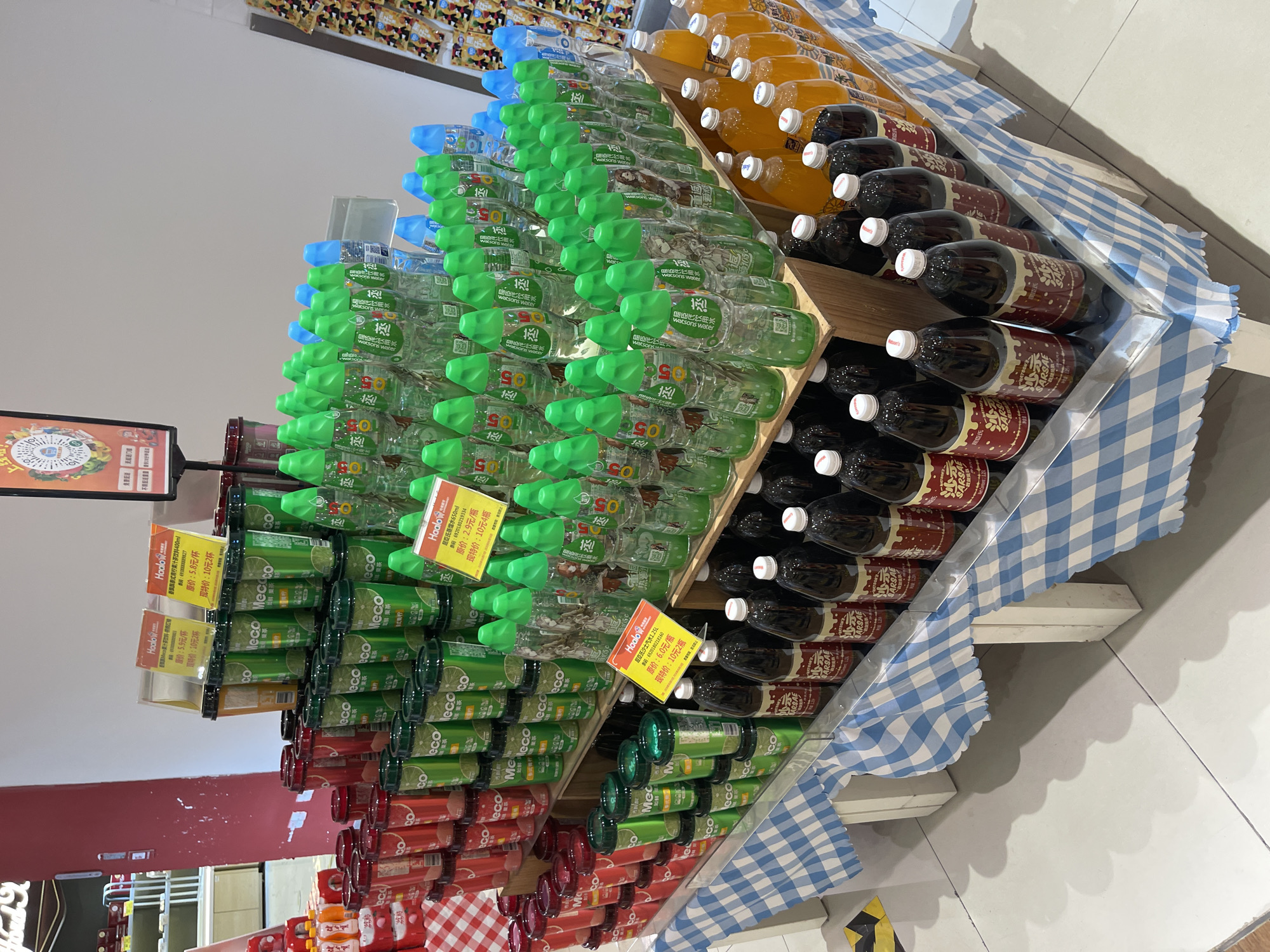 The image shows a retail display of beverages in what appears to be a supermarket aisle in Panyu District, China. The display is tiered, with various bottles and cans of drinks arranged on wooden shelves covered with a blue and white checkered tablecloth.
Dominating the display are rows of green plastic bottles with green caps, some with white labels featuring Chinese characters and images. To the left, there are stacks of red and green cans, also with Chinese labeling. Further to the right, there are clear plastic bottles with green and blue caps. In the foreground, there are dark-colored bottles, likely sodas, with red and white labels and white caps. Behind these, there are rows of orange-colored beverages in plastic bottles.
Several yellow price tags with Chinese text are visible, indicating promotional pricing. The background is a plain white wall, with a hint of shelving and product packaging to the far left. The lighting suggests an indoor commercial setting. No people are visible in the frame.