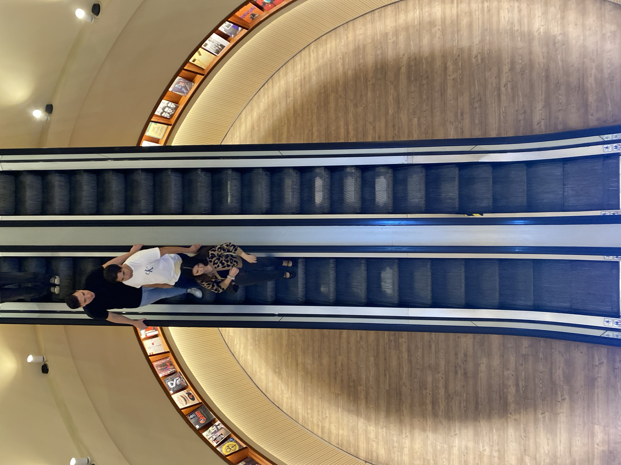 An aerial interior shot depicts four individuals descending a multi-level bookstore on an escalator in El Ateneo Grand Splendid, Santa Fe Avenue 1860, Recoleta, Buenos Aires, Argentina. Two parallel escalators with dark grey steps and black handrails are centrally positioned, leading downwards to a curved, light-toned wooden floor. Along the curved inner wall, a recessed wooden display shelf exhibits numerous books or album covers, illuminated by a continuous strip light. The upper wall and ceiling are a light cream color, featuring several circular recessed light fixtures.
On the left escalator, from top to bottom:
1. An individual's lower legs and feet, clad in dark pants and shoes, are partially visible.
2. A male in a black t-shirt and light blue jeans stands, his right hand on the handrail.
3. A male in a white t-shirt with a "CK" logo and light pants stands in front, his left hand on the handrail.
4. A female in a leopard-print long-sleeved top, dark pants, and dark flat shoes stands alone further down the escalator, looking forward.
All visible subjects are passively riding the escalator downwards.