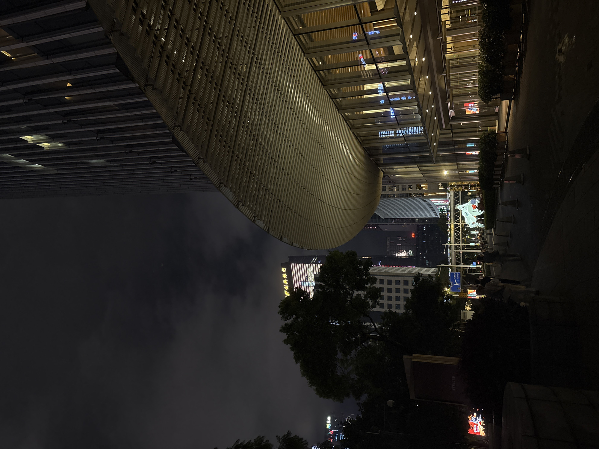 This is a nighttime, outdoor, low-angle shot of a modern urban scene in Tianhe District, China. The image is vertically oriented. In the foreground, a curved, ribbed structure, likely part of a building's overhang or an architectural element, dominates the upper left portion. Beneath it, illuminated windows of a glass-fronted building reflect city lights. To the right, a paved walkway lined with potted plants and bollards leads into the distance.
Further back, several tall buildings are visible. One building in the middle distance features illuminated Chinese characters, possibly a brand name or business. Another building has a grid-like facade. In the far distance, the city skyline is dotted with lights. A large, white, abstract sculpture, possibly an animal, is visible on the right side of the image, with several people gathered around it, suggesting a public space or event.
The sky is dark and cloudy, indicating it is nighttime. The overall atmosphere is one of a busy, well-lit urban environment at night, with a focus on modern architecture and public spaces. The presence of the sculpture and gathering people suggests a potential public art installation or festive display.