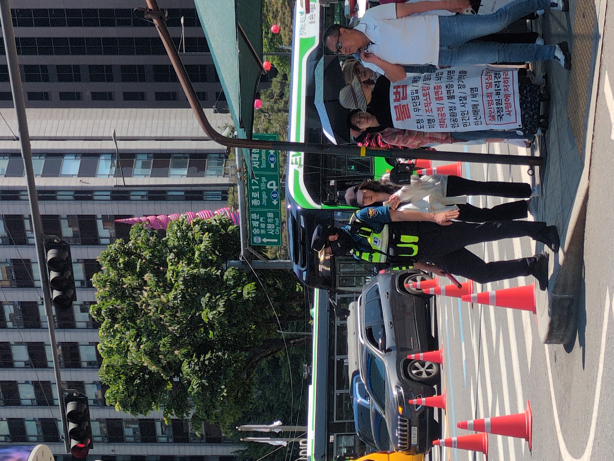 The image shows a street scene in Seoul, South Korea. A group of people are standing on a sidewalk, holding signs with Korean text, indicating a protest or demonstration. An officer in a safety vest stands behind them. A bus is parked nearby, possibly related to the demonstration. Orange traffic cones line the street near parked vehicles. Street signs show the locations 숭례문 (Sungnyemun), 종로1가 (Jongno 1(il)-ga), and 서대문 (Seodaemun), which are districts within Seoul. The signs also indicate the direction to the 시청 (City Hall). A large tree and building are visible.