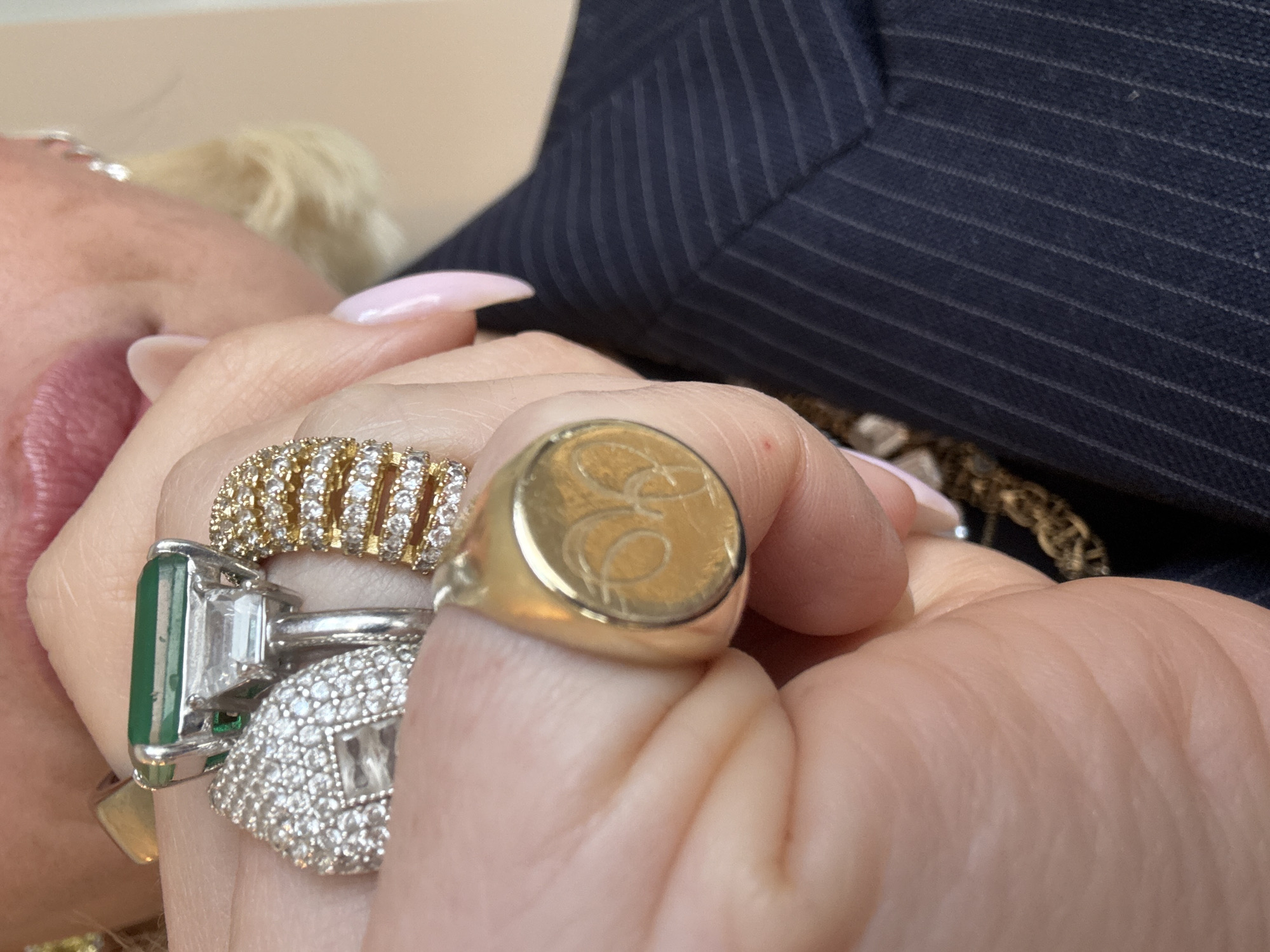 A close-up image focuses on the left hand of an individual, which is partially clenched, prominently displaying several rings. On the ring finger, an emerald-cut green gemstone ring with flanking clear stones is visible, alongside a pave-set white metal ring featuring numerous small clear gemstones. A wide gold band ring, also pave-set with small clear gemstones, is present. On the index finger, a gold signet ring with an engraved oval face, depicting a monogram or crest, is worn. The fingernails are manicured with a light pink polish. In the blurred background, the individual's lips with pink lipstick, blonde hair, and a diamond earring are discernible. A dark navy blue pinstripe suit jacket is visible on the right side of the frame, underneath which a gold chain is present. The environment appears to be an indoor setting with a plain, light-colored wall as the background. No specific location within London, United Kingdom, is identifiable from the visible elements.