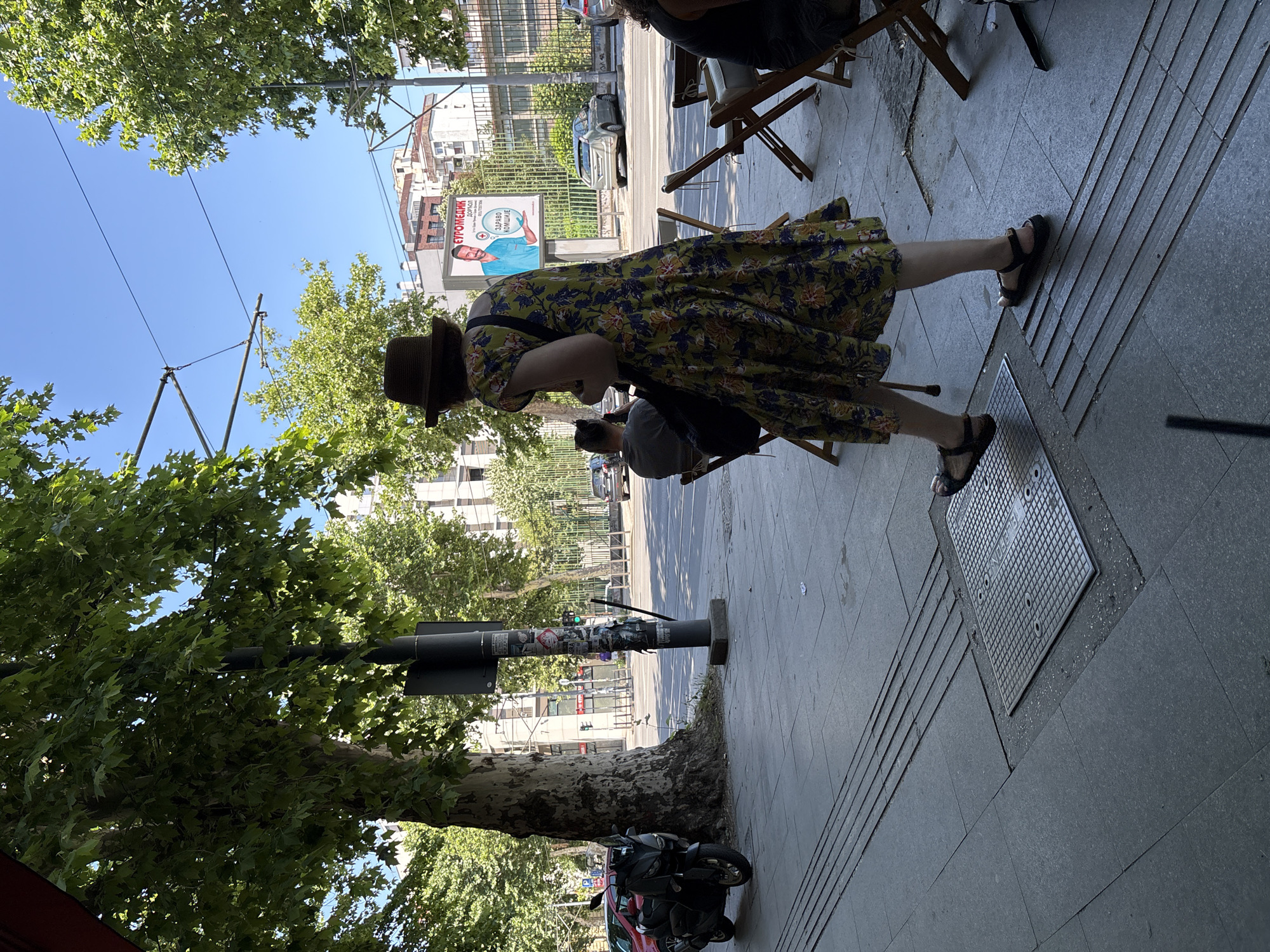 The image shows a woman walking on a sidewalk in Belgrade, Serbia. She is wearing a floral dress and a hat. In the background, there are trees, buildings, and a billboard with an advertisement. The billboard shows a man in a blue medical uniform and text in Cyrillic script. The scene appears to be a sunny day, with a clear blue sky. There are a few parked cars and a scooter visible on the street. There are also tables and chairs set up on the sidewalk, suggesting an outdoor seating area for a cafe or restaurant. A man is seated at one of these tables in the background. There is also a metal manhole cover on the sidewalk.