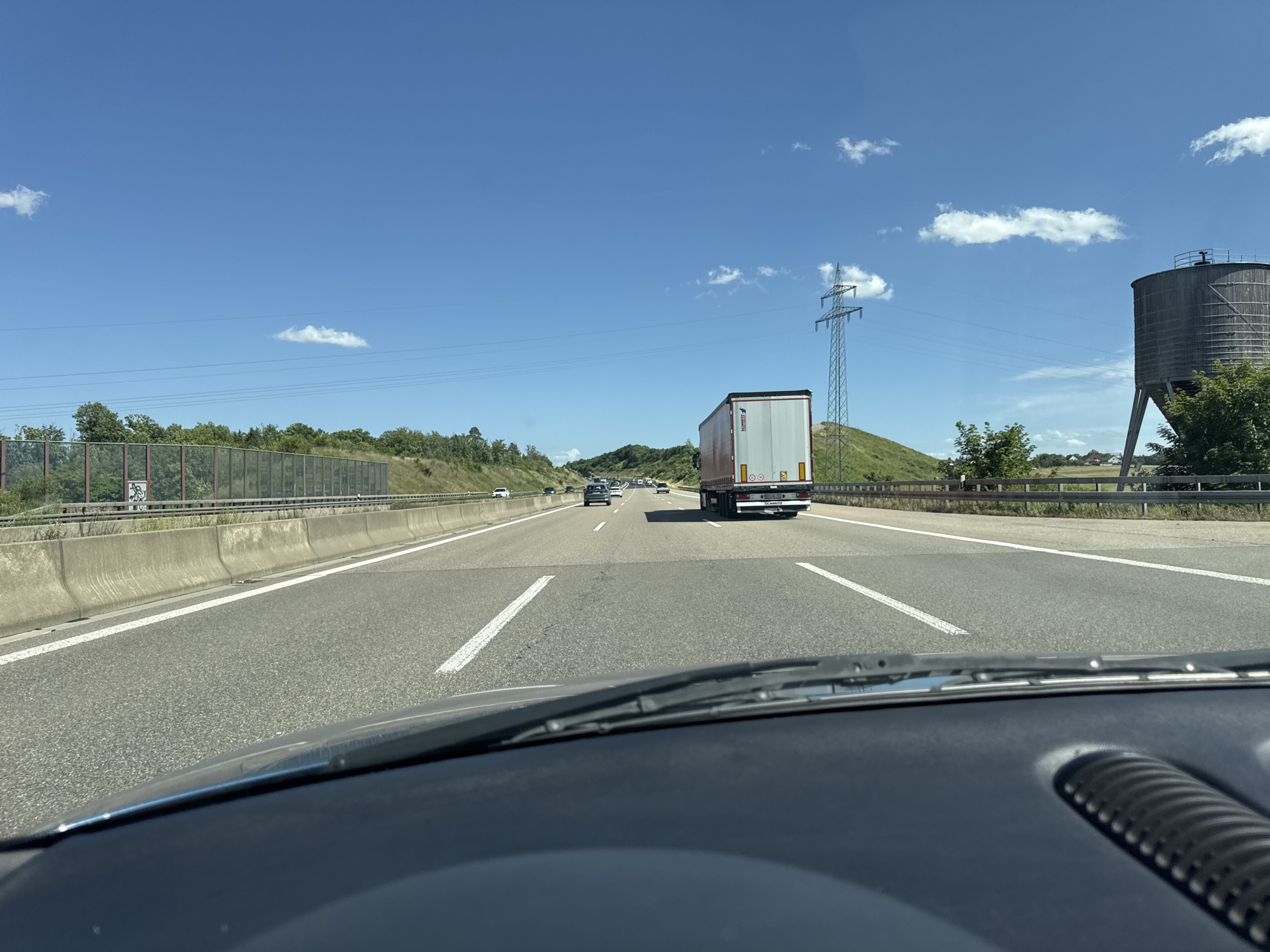 The image shows a view from the inside of a car traveling on a highway. The highway has multiple lanes and is bordered by concrete barriers and green hills with trees. A large semi-trailer truck is visible in the right lane, ahead of the car. The sky is blue with scattered white clouds, and a tall electrical pylon is visible on the right side of the road. In the distance, more cars and trucks can be seen on the highway. A large, cylindrical silo structure is also present on the right side of the highway, behind the electrical pylon. The car's dashboard and windshield wipers are in the foreground. This scene is located on the A8 Autobahn near Wimsheim, Germany.