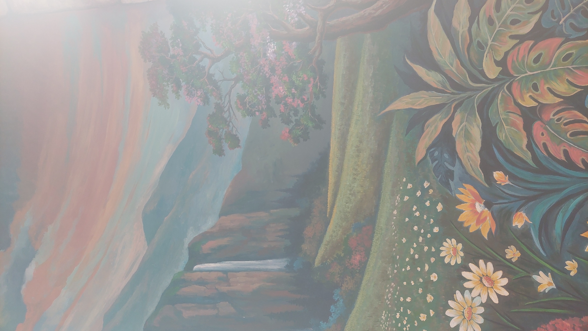 This is a mural painted on a wall, depicting a serene landscape. The scene features a towering waterfall cascading down a rocky cliff on the left side, surrounded by lush green trees and rolling hills. In the mid-ground, a flowering tree with pink and white blossoms adds a splash of color. The foreground is filled with a field of small white flowers, with larger, brightly colored flowers, including yellow and orange ones, closer to the viewer. Large, tropical-looking leaves in shades of green and orange are prominent in the bottom right corner. The sky is painted with streaks of pink, orange, and blue, suggesting a sunrise or sunset. The overall impression is one of natural beauty and tranquility. The mural appears to be located outdoors, possibly on the exterior wall of a building in Cileungsi, Indonesia. There are no people or animals depicted in the mural.