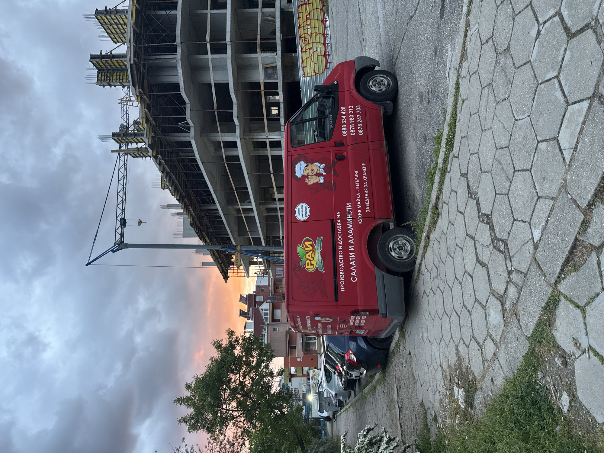 The image depicts a street scene in Blagoevgrad, Bulgaria. A red commercial van with the name "Рай" and Cyrillic text advertising salads, catering, and food establishments is parked on the side of the street. Several other parked cars are behind the van. A construction site with a tall concrete building under construction and a crane are visible in the background, and the sky shows shades of pink and grey, suggesting either sunrise or sunset. The sidewalk is paved with hexagonal concrete blocks.