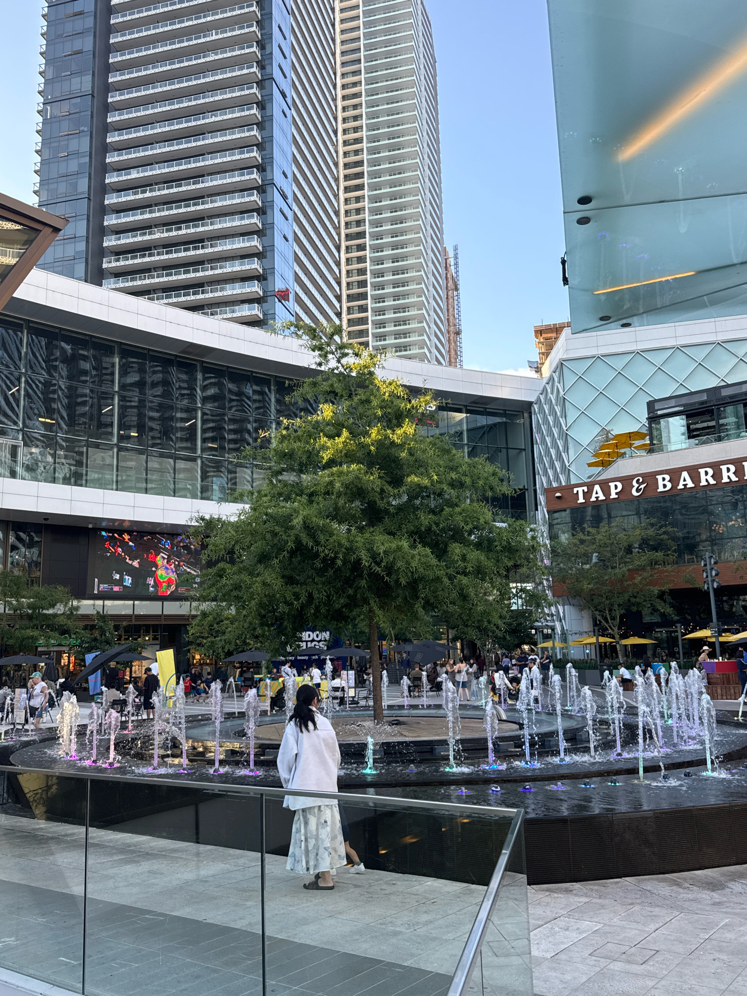 This image depicts a modern urban plaza in Burnaby, Canada, featuring contemporary architecture and public amenities. Dominating the background are two towering, glass-clad skyscrapers, one with distinct horizontal balconies and the other featuring a more uniform facade. In the mid-ground, a multi-story building with large glass windows displays a vibrant LED screen showing a colorful, abstract image. Adjacent to this is another building with the signage "TAP & BARREL". The foreground is occupied by a decorative fountain with multiple water jets illuminated with colored lights, creating a dynamic visual effect. Several people are visible in the plaza; one person in the foreground, facing away from the camera, wears a white jacket and a patterned skirt. Other individuals are scattered throughout the plaza, some seated at tables under umbrellas, others walking, and a group is gathered near the LED screen. The environment appears to be a lively public space designed for recreation and commerce, with trees and seating areas contributing to the ambiance. The sky is clear and blue, suggesting daytime.