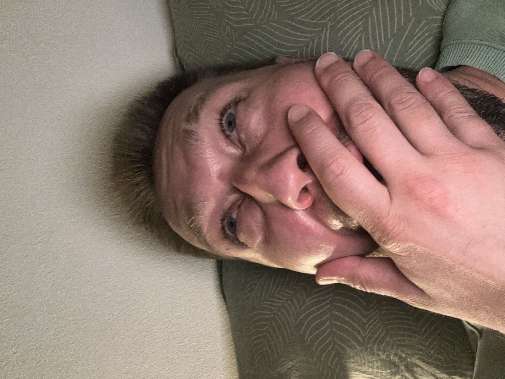 A close-up, rotated portrait shows a person lying down, their face obscured by their own hand. The person's left eye is visible, open and looking towards the camera, with a blue iris and dark eyelashes. The skin on their face appears flushed, possibly from lying down or exertion. The hand covering the face is placed over the nose and mouth, with the fingers splayed across the lower half of the face. The texture of the skin on the hand is detailed, showing creases and pores. The person has light brown hair.
The background consists of two distinct textures. To the left is a plain, light-colored surface, possibly a wall or bedding, with a subtle grainy texture. To the right, a darker green fabric with a repeating pattern of thin, white, wavy lines is visible, likely a piece of clothing or upholstery. A hint of a ribbed collar, suggesting a sweater or sweatshirt, is visible in the upper right corner.
The image appears to be taken indoors, in a dimly lit environment, suggested by the soft lighting and shadows. There is no visible text. The overall mood conveyed is one of introspection, exhaustion, or perhaps discomfort, with the hand shielding the face from view. The orientation of the image suggests it was taken by the subject themselves, possibly in a relaxed or reclined position.