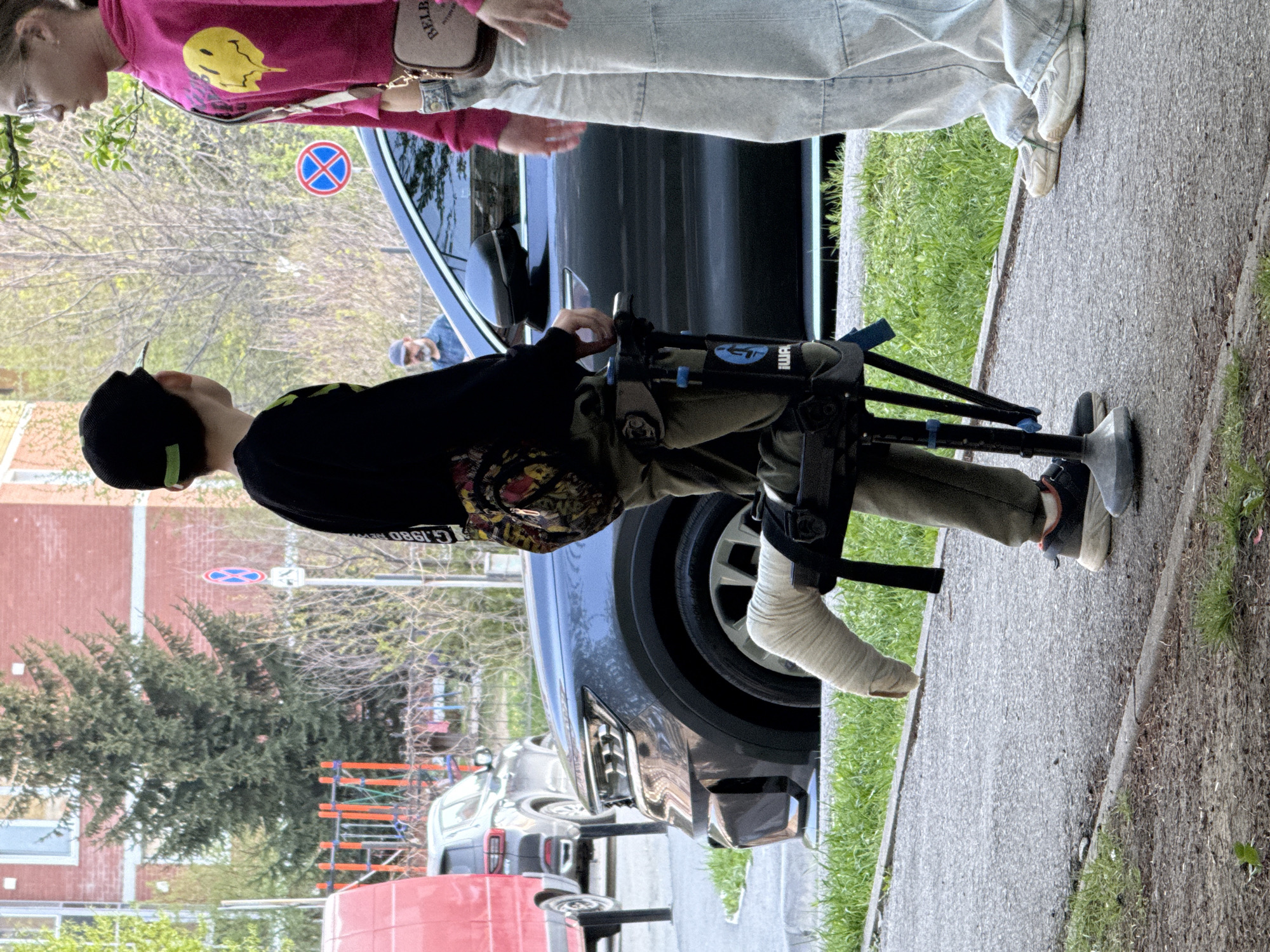 A full shot shows a person with a cast on their left leg standing in a knee walker, near a parked dark-colored car. A person in a pink top stands to the right. The setting is an urban street in Yekaterinburg, Russia, based on the road signs. Trees, buildings, and other parked cars can be seen in the background.