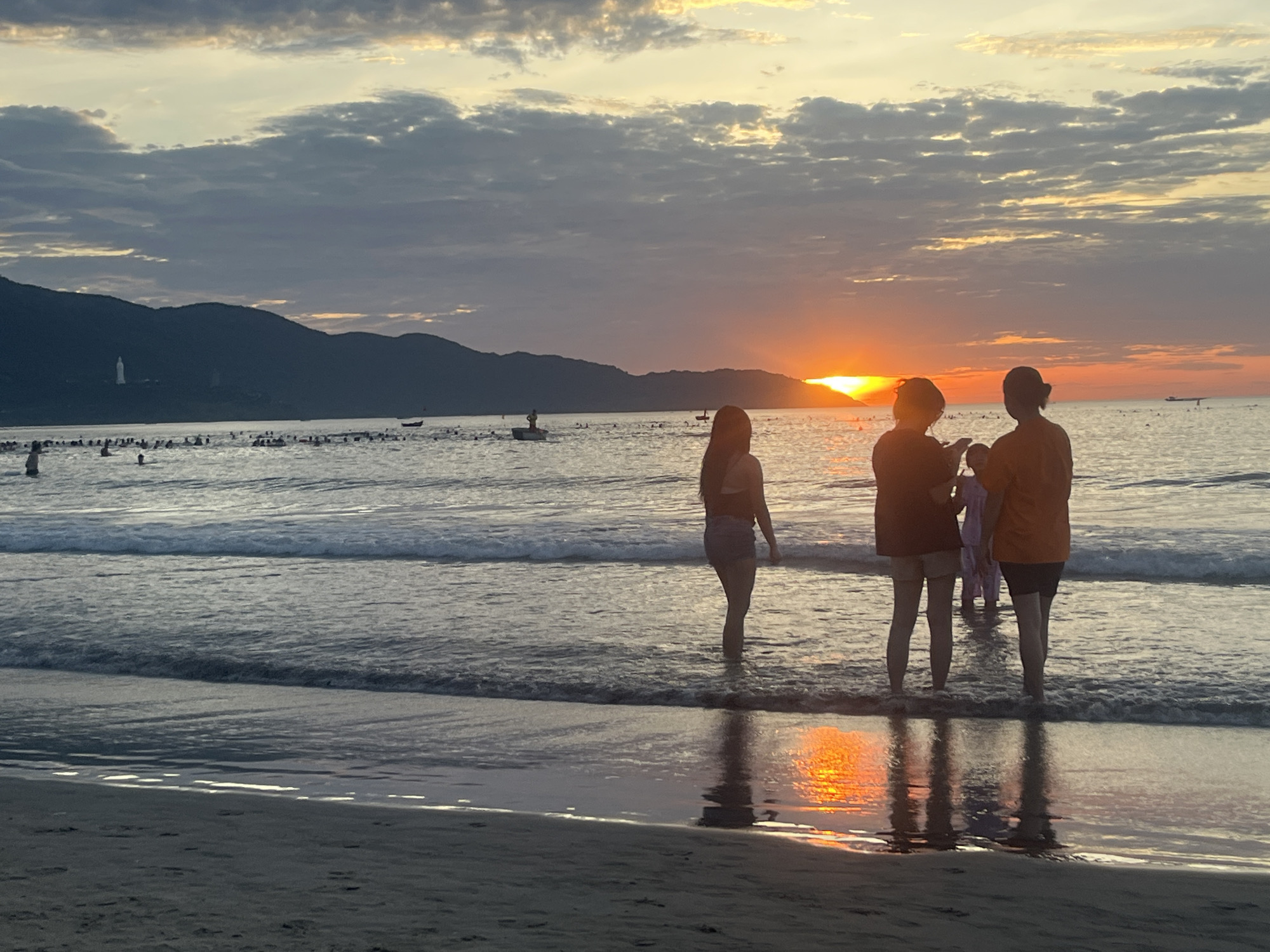 This image captures a sunset scene on a beach in Đà Nẵng, Vietnam, with a perspective looking approximately northwest. The sun, a prominent orange-red disc, is partially obscured by the distant horizon on the right, casting a strong, elongated reflection of orange and yellow light across the water and wet sand towards the viewer.
The sky above features scattered, dark grey clouds against a lighter grey-yellow background, transitioning into vibrant orange and yellow hues near the horizon. To the far left, a distinct white statue, identified as the Lady Buddha (Goddess of Mercy) of Linh Ung Pagoda, is visible on the highest peak of a distant, dark mountain range, likely the Son Tra Peninsula.
The foreground consists of dark, wet sand with gentle waves breaking at the shoreline, displaying white foam. Four individuals are positioned in the shallow water directly in front of the camera, appearing as dark silhouettes against the sun's reflection. From left to right, one person in shorts and a top; two people grouped together, one appearing to hold a small child; and a fourth individual. Their reflections are clearly visible on the water.
Mid-ground details include numerous individuals dispersed across the ocean, wading or swimming, visible as dark forms. Several small boats are also scattered across the water, with at least one boat containing a standing person. The overall scene depicts a populous beach at dusk.