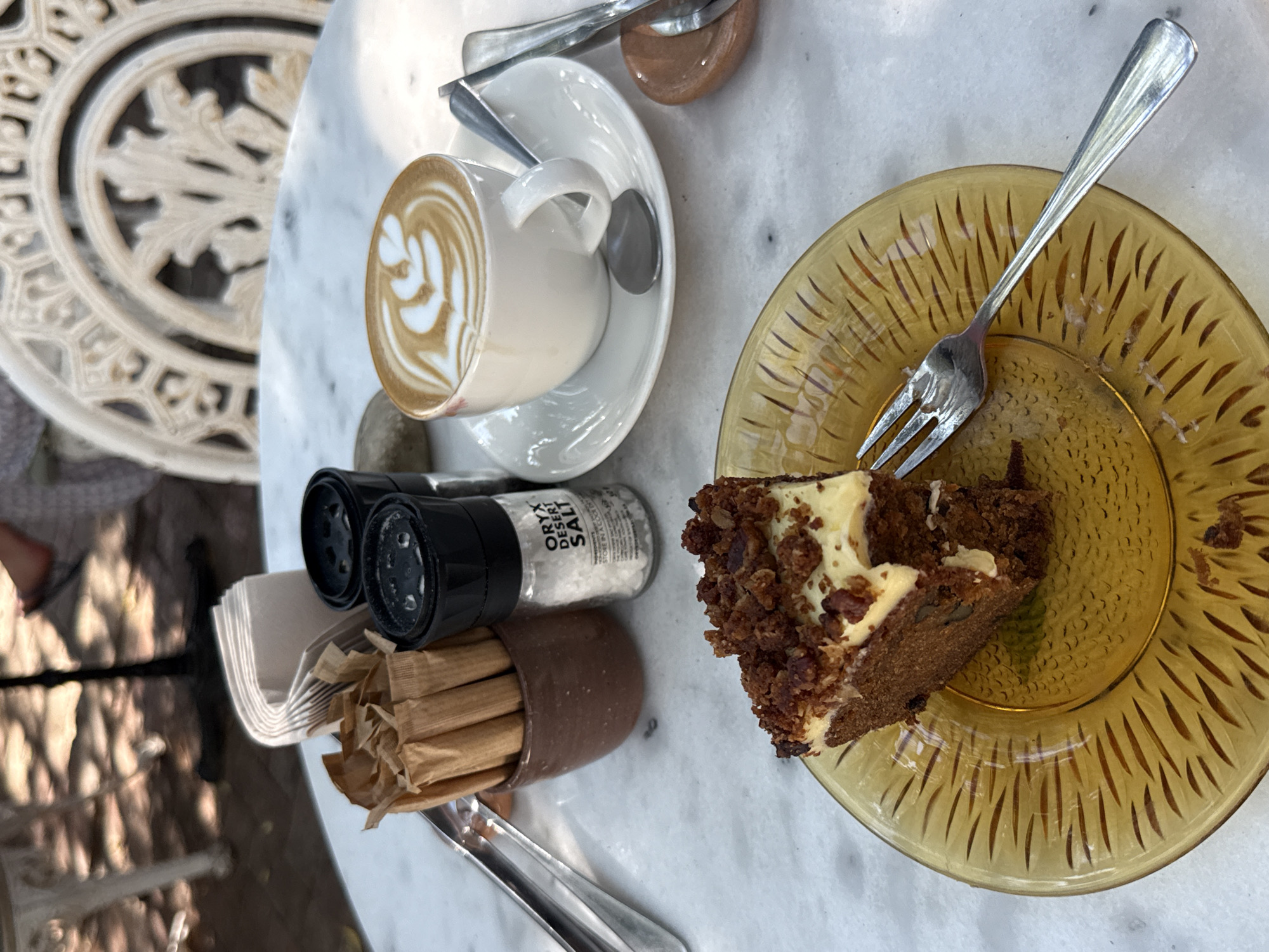 The image shows a partially eaten slice of carrot cake on a textured amber glass plate, along with a latte in a white cup and saucer on a marble table. A fork rests on the plate. A brown ceramic container filled with sugar packets and a salt shaker labeled "ORYX DESERT SALT" are also on the table. Part of an ornate metal table is visible in the background. Based on the salt and likely the style of the café, the location is probably in Cape Town, South Africa.