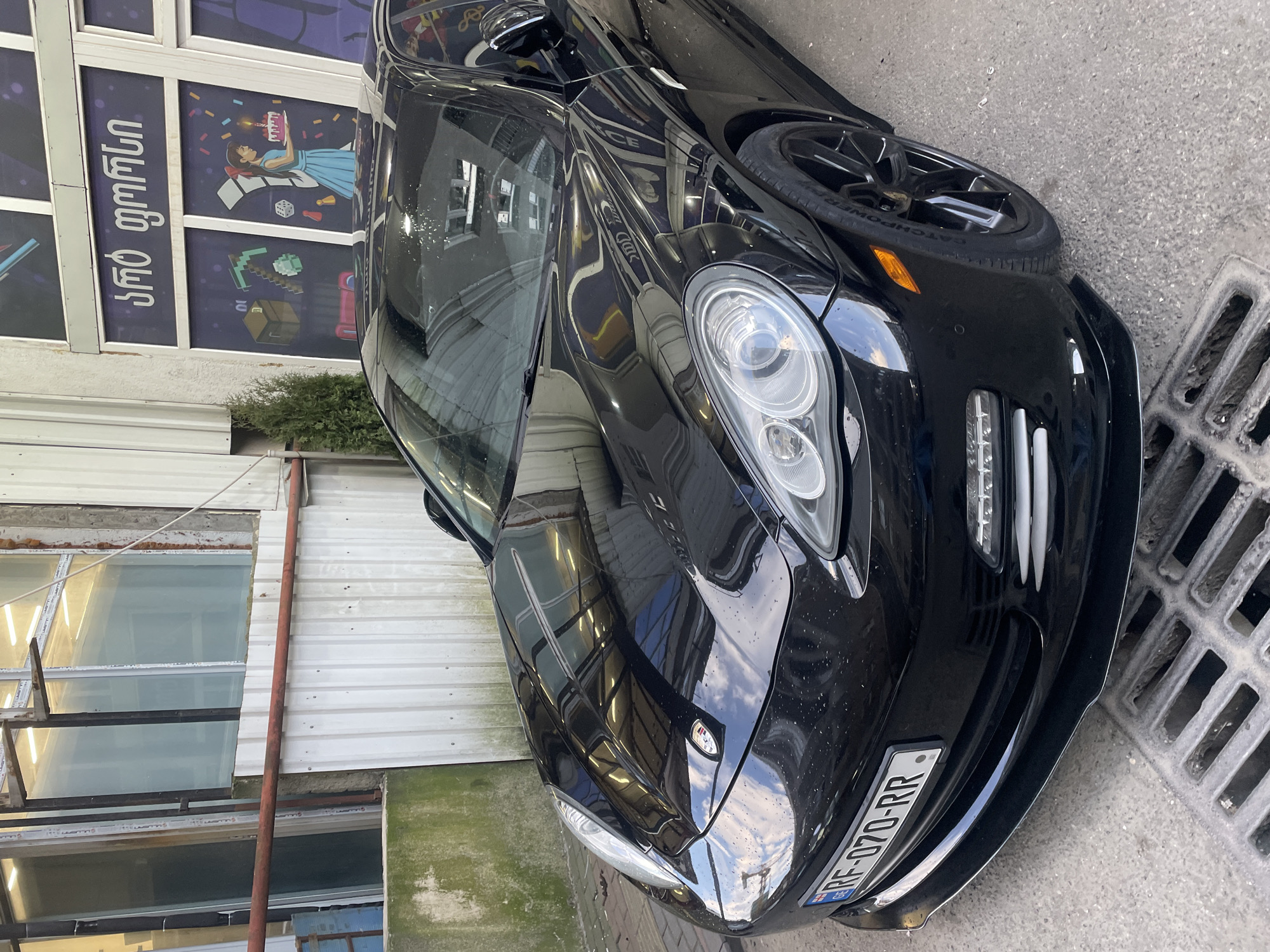 A close-up shot depicts the front of a black Porsche Panamera, parked at an angle on a street in Batumi, Georgia. The car's headlights, front bumper, and license plate ("RF-070-RR") are visible. The reflection of the street scene is visible on the car's hood. Adjacent to the car is a concrete drain. In the background, a building with windows and signage featuring Georgian script and cartoon illustrations (including a Minecraft-style block) is visible. The area appears to be an urban setting with buildings of varying conditions.