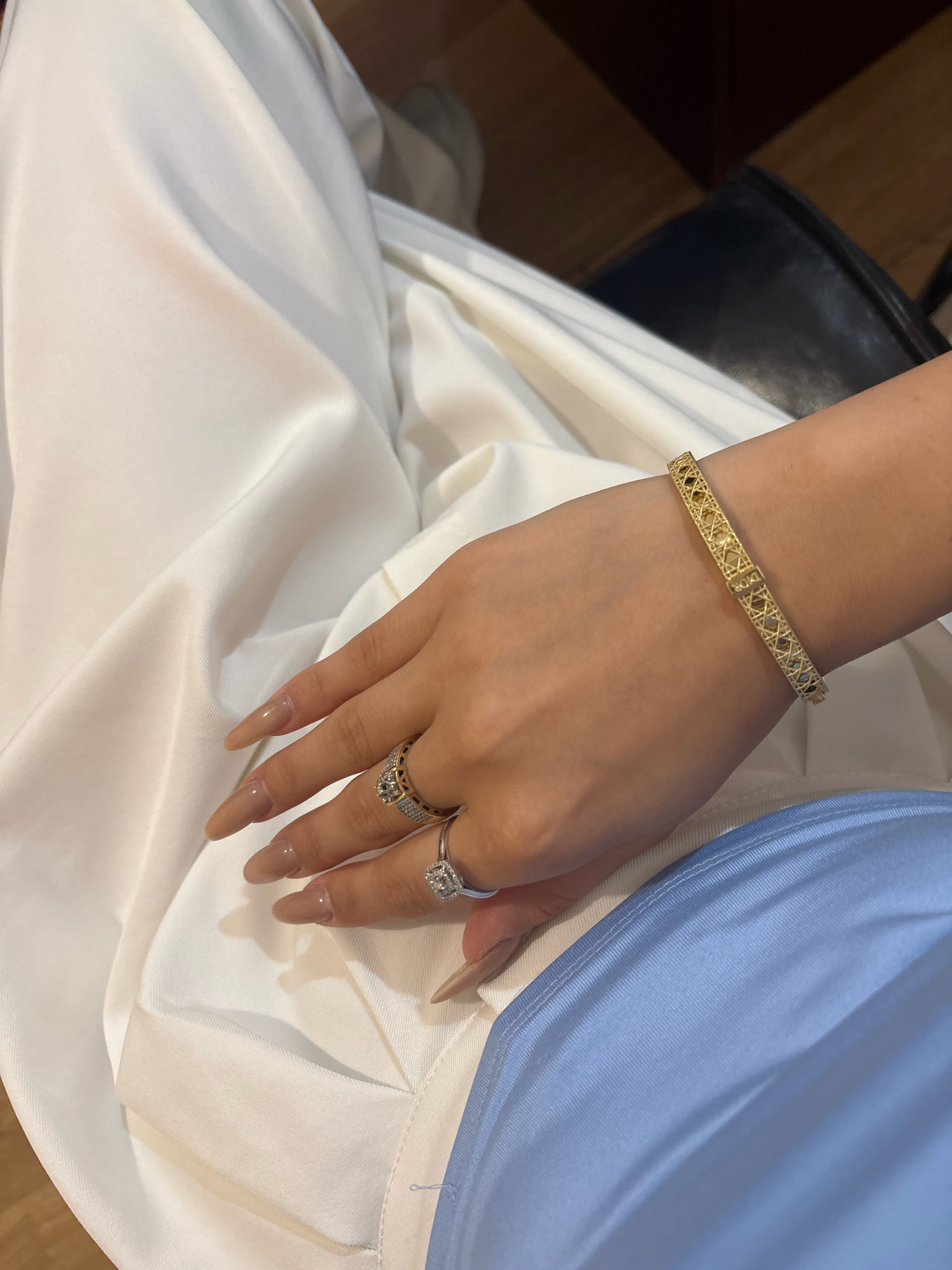 The image is a close-up shot of a person's hand and arm, adorned with jewelry, resting on a white fabric. The hand has long, almond-shaped nails painted in a neutral tone. Two rings are visible on the ring finger: a larger one with a prominent square-cut stone surrounded by smaller stones, and another band-style ring with intricate detailing. On the wrist, a thin gold bracelet with a geometric, openwork design is worn. The person is wearing a light blue garment on their lower body. The background is blurred, showing hints of dark furniture and wooden flooring, suggesting an indoor setting. There is no indication of the city of Ijevan, Armenia, in the image.